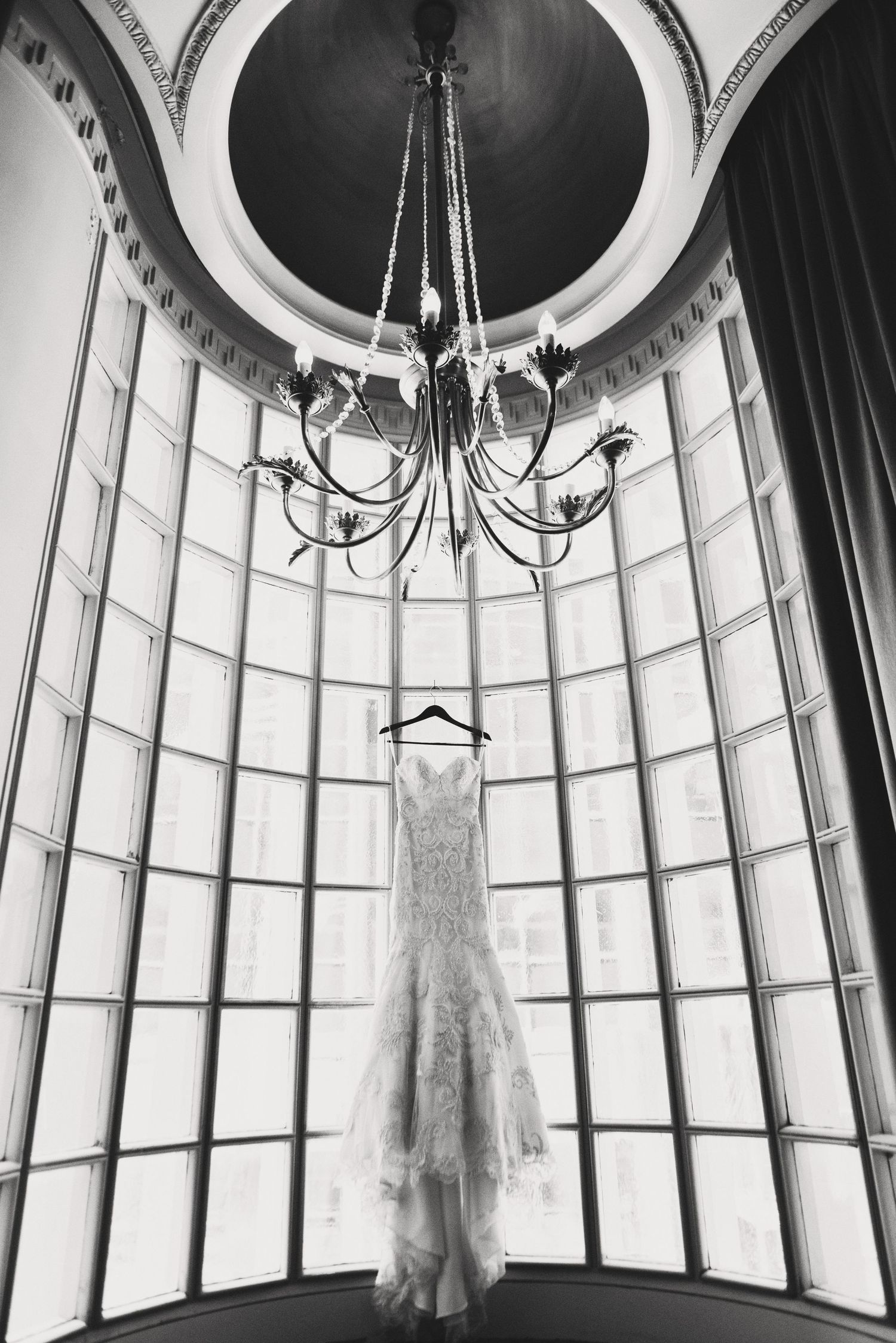 A white lace wedding dress hangs elegantly in a curved window alcove beneath an ornate chandelier in black and white. Athletic Club