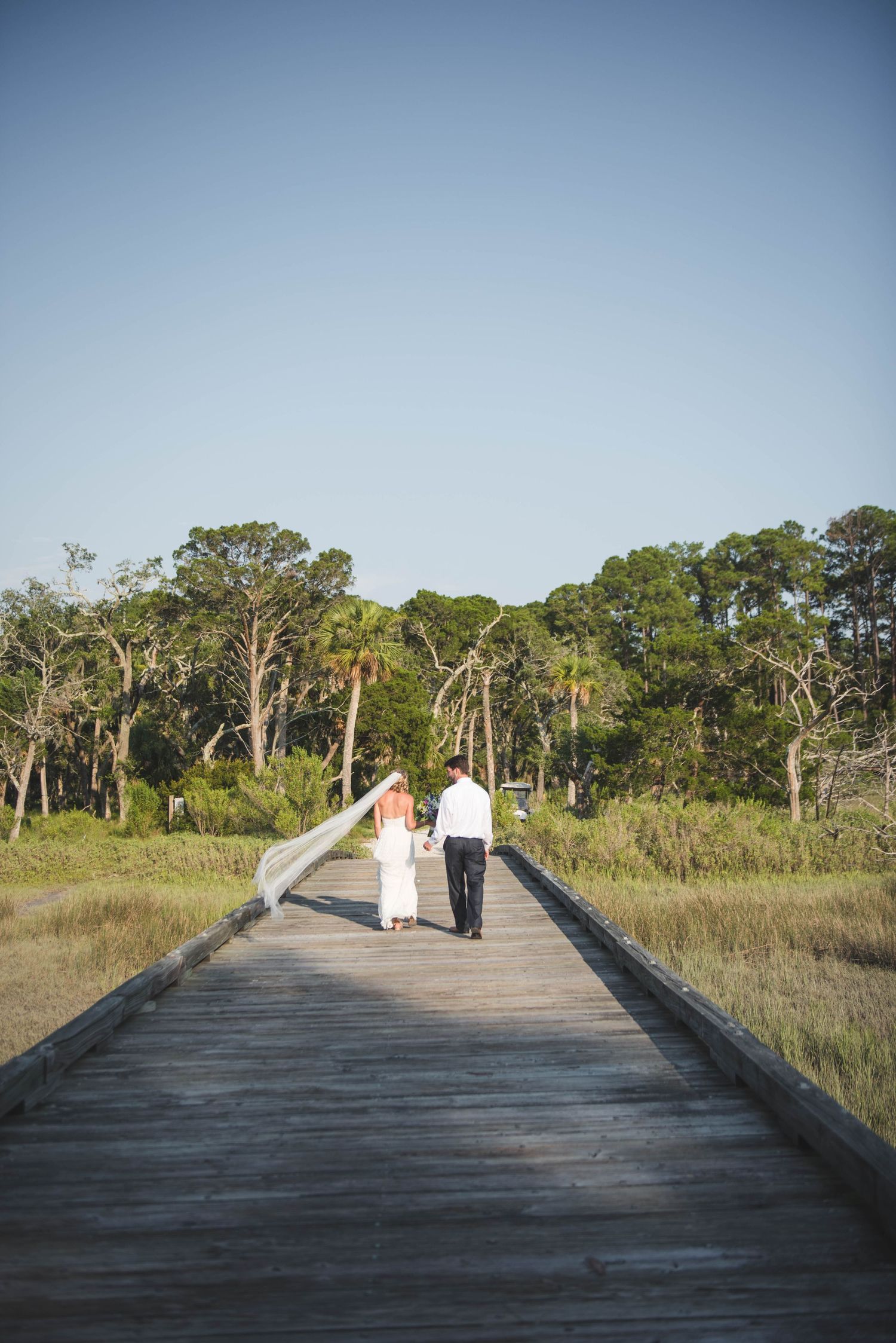 Bride and groom walk hand-in-hand down a wooden boardwalk surrounded by lush greenery on their wedding day.