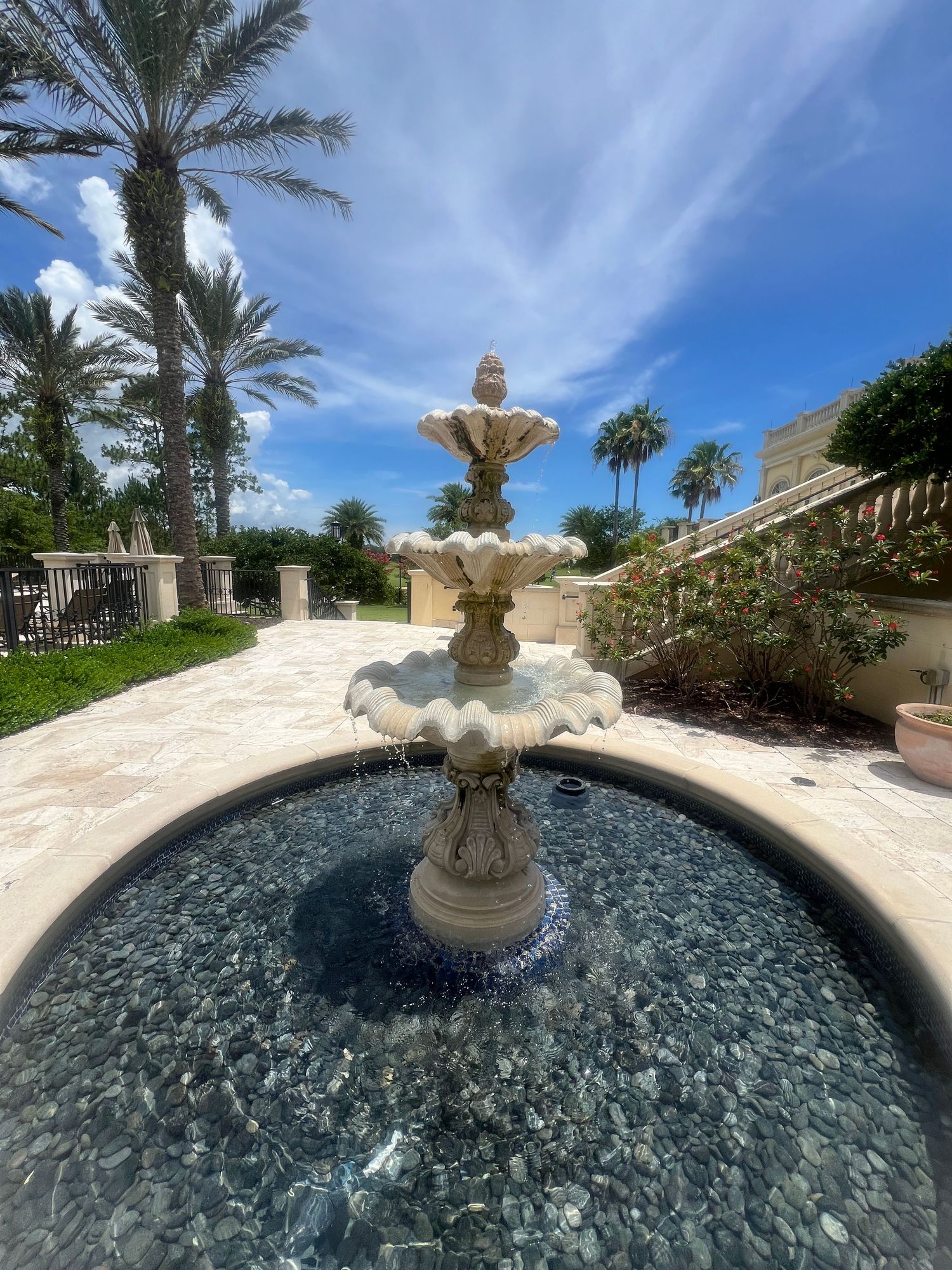 Three-tier ornate stone fountain surrounded by palm trees and travertine pavers under a blue sky.