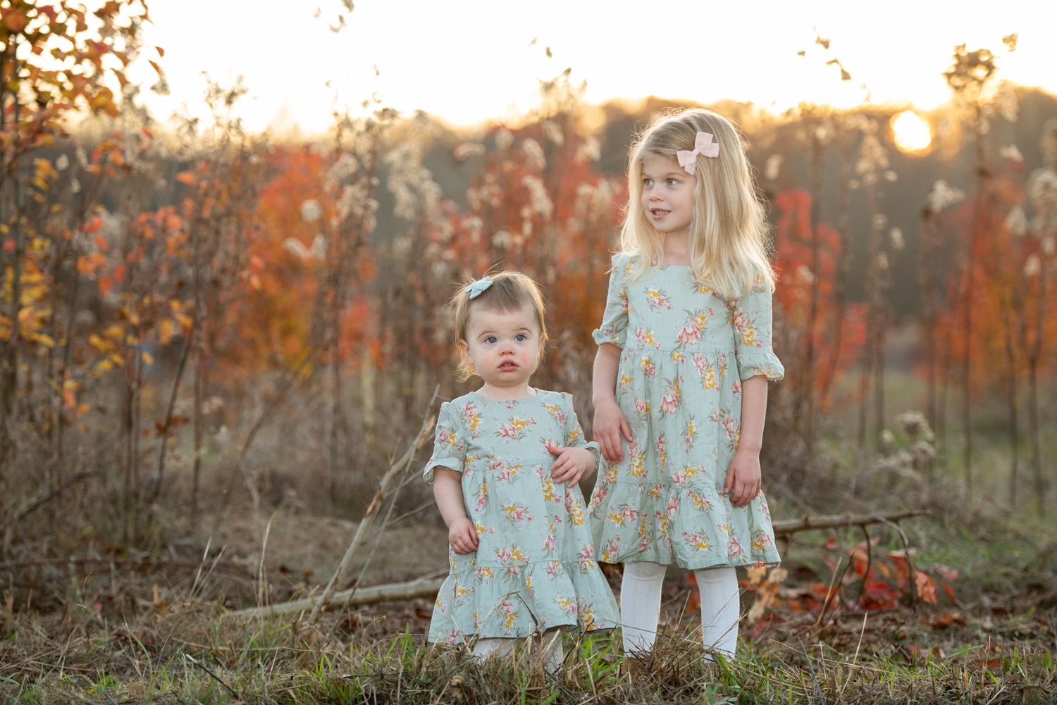 A group in matching blue floral dresses stands together in a sunlit autumn field with orange foliage in the background.
