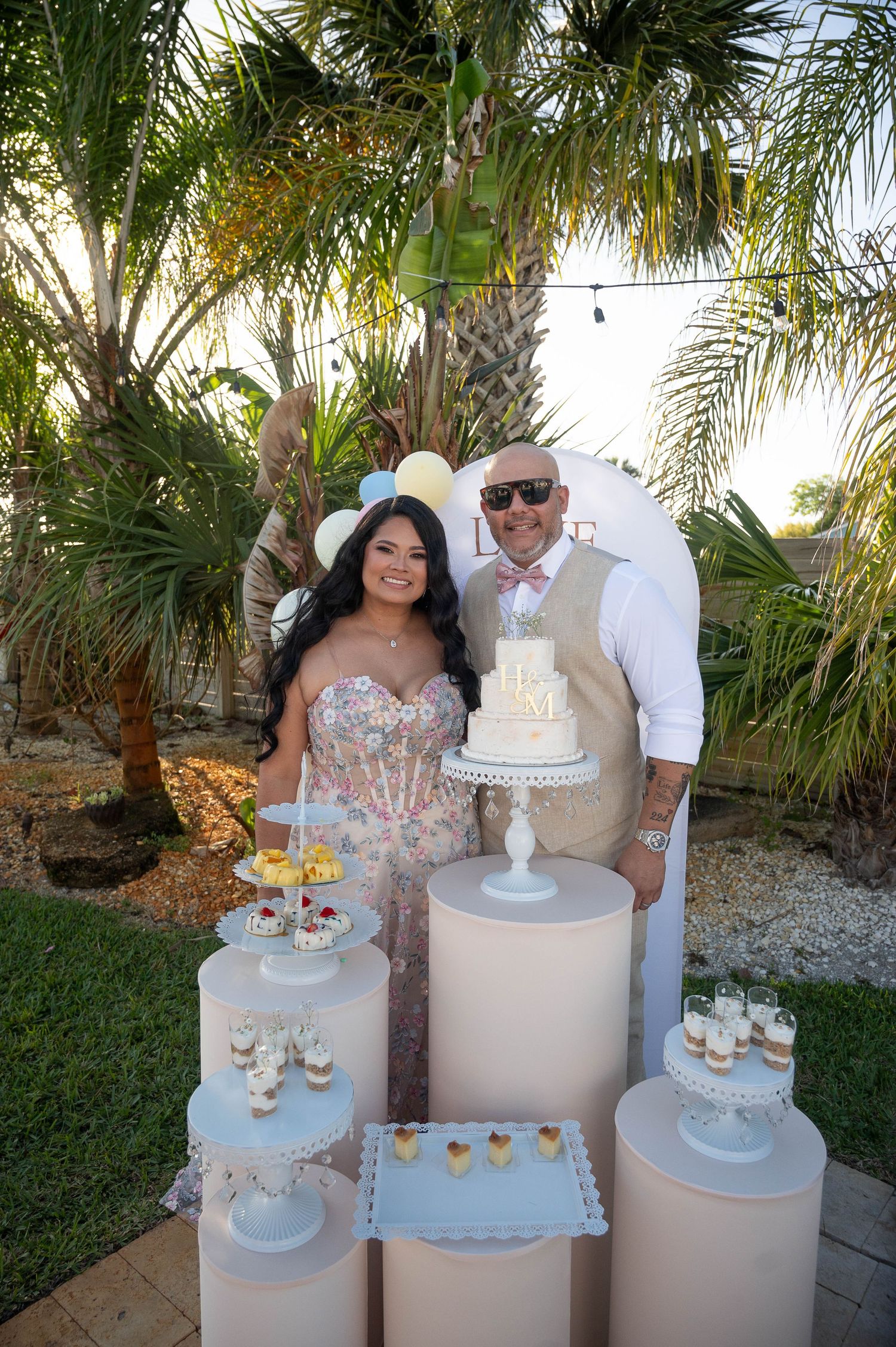Couple posing with elegant white wedding cake and dessert display outdoors among tropical palm trees.