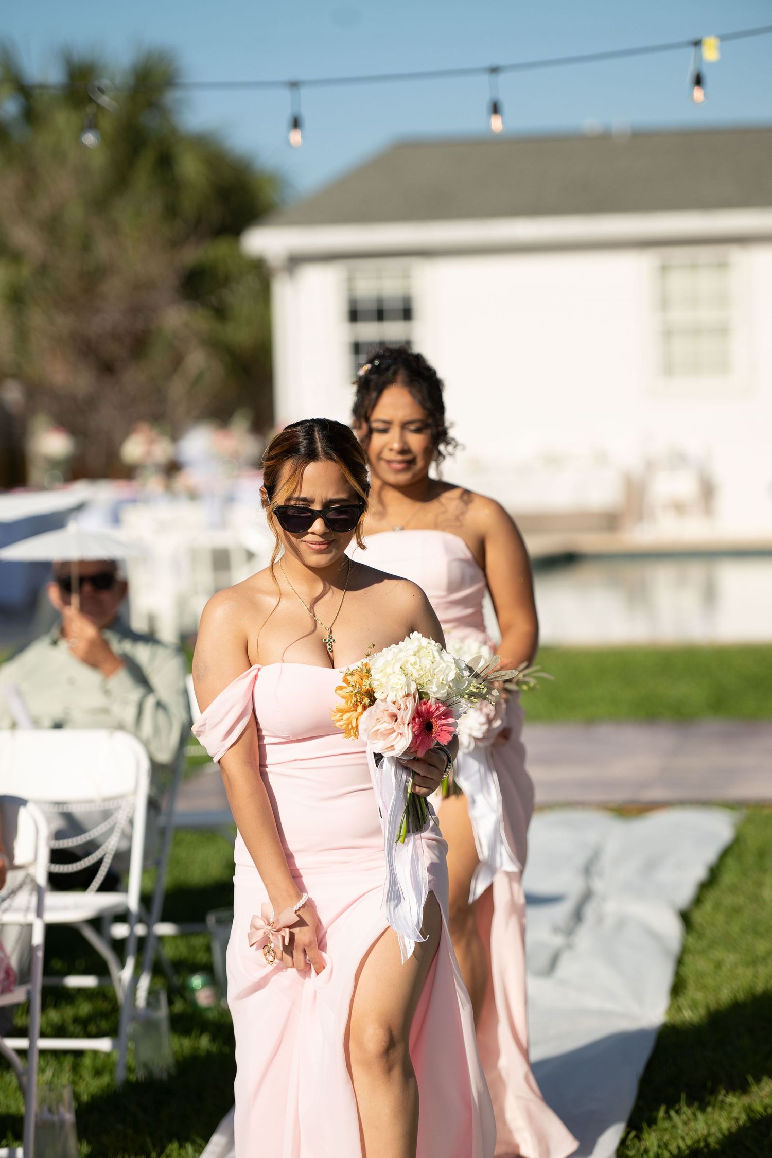 Two bridesmaids in pink off-shoulder dresses walk down outdoor wedding aisle, one holding a colorful bouquet.