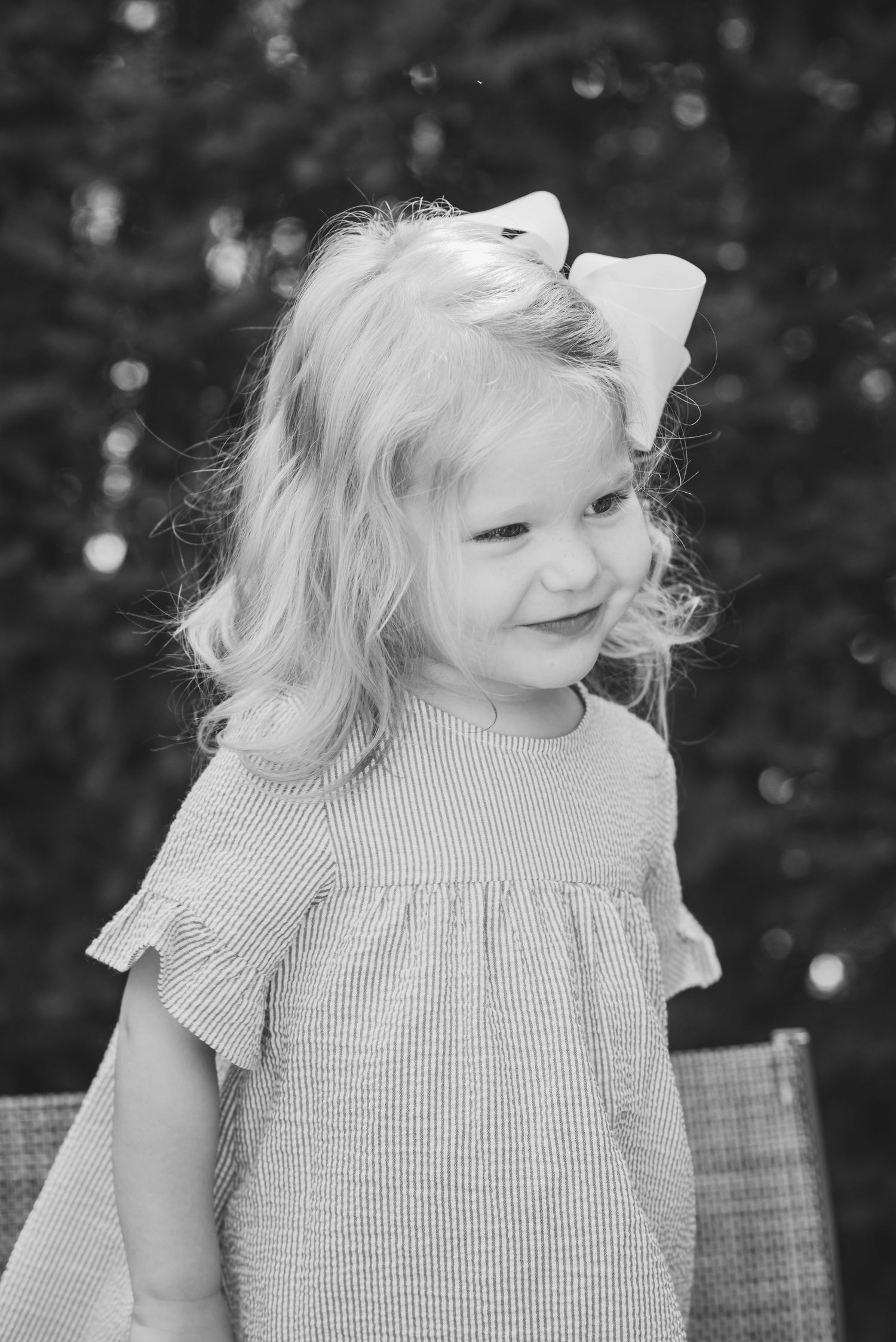 Black and white portrait of a young blonde girl wearing a bow and ruffled dress, smiling outdoors.
