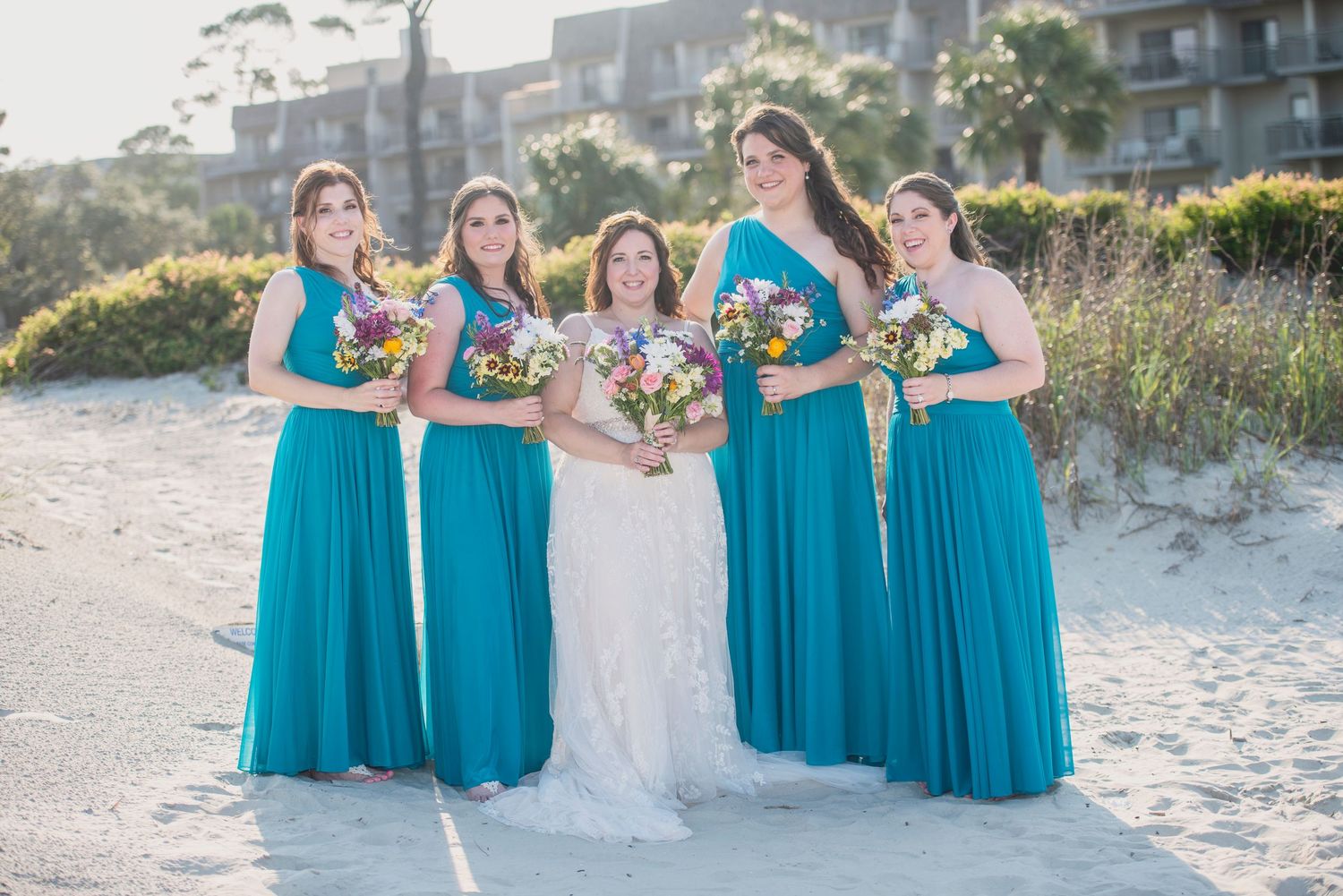 Wedding party in turquoise bridesmaid dresses and white bridal gown pose on beach.