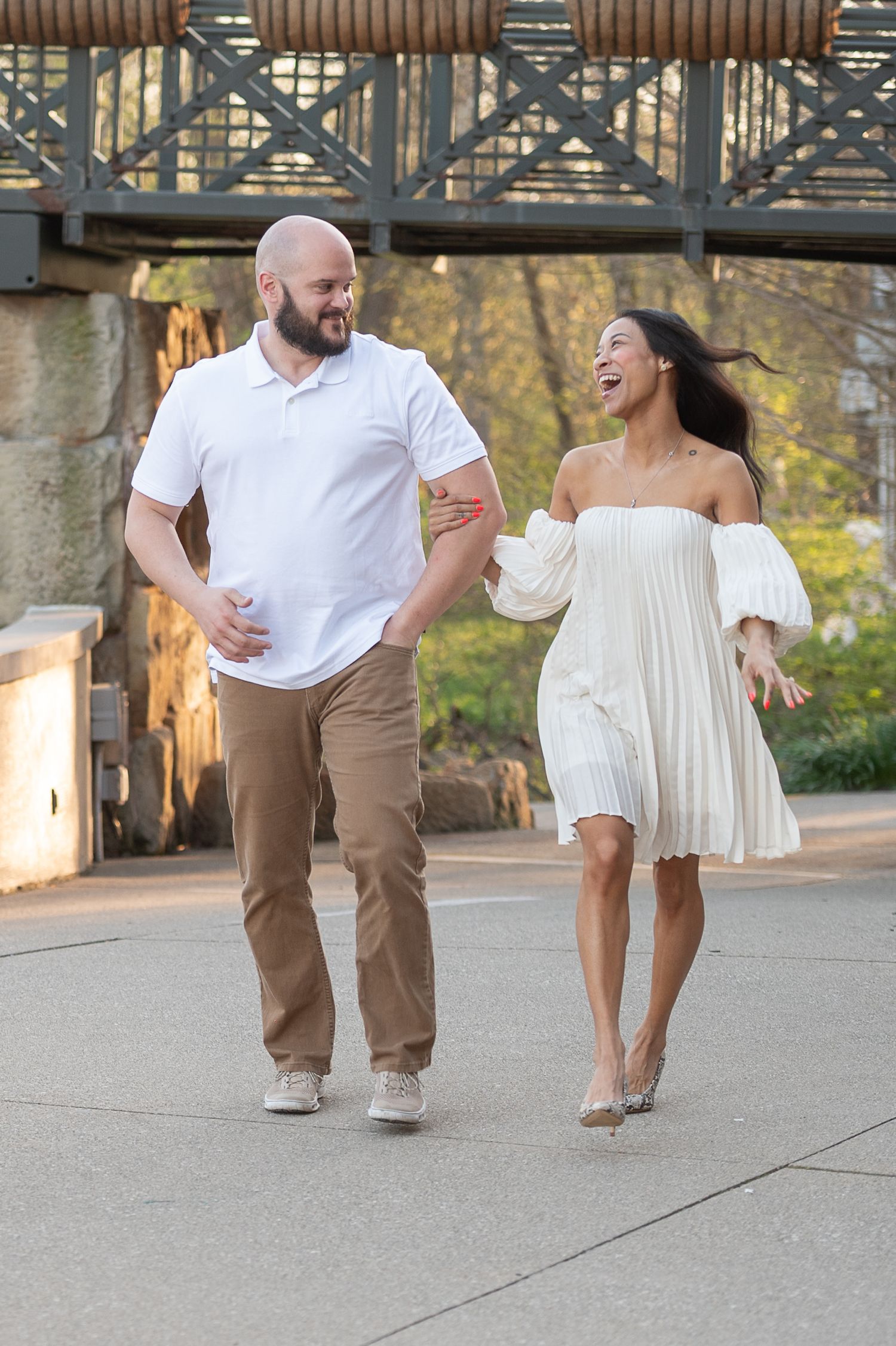 Playful couple in casual outfits sharing laughs while walking under bridge in park setting. Hammock Beach engagement session.