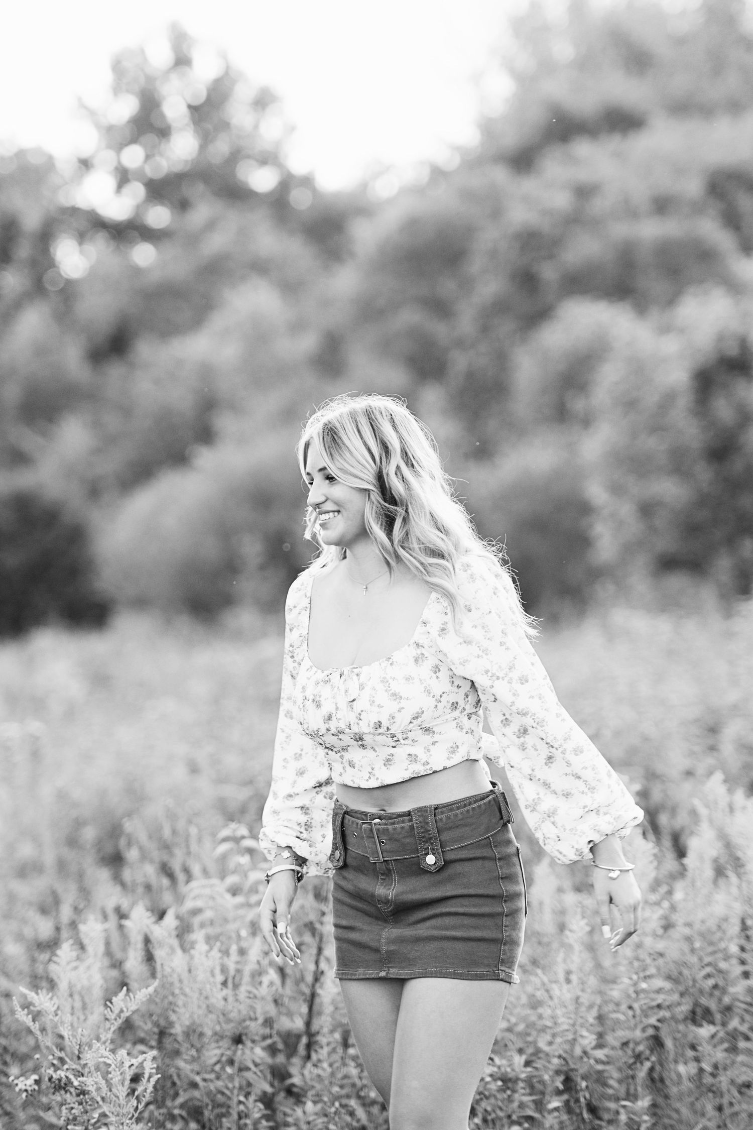 Black and white photo of a smiling blonde woman in a floral top and denim skirt standing in a field.
