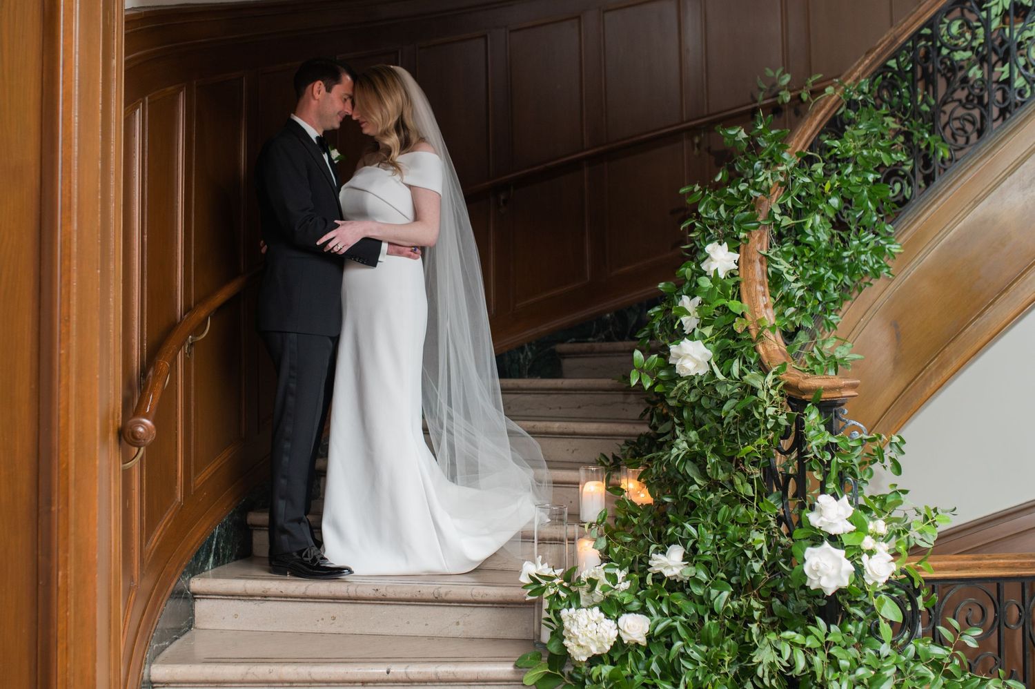 Wedding party descends an elegant wooden staircase adorned with floral decorations.