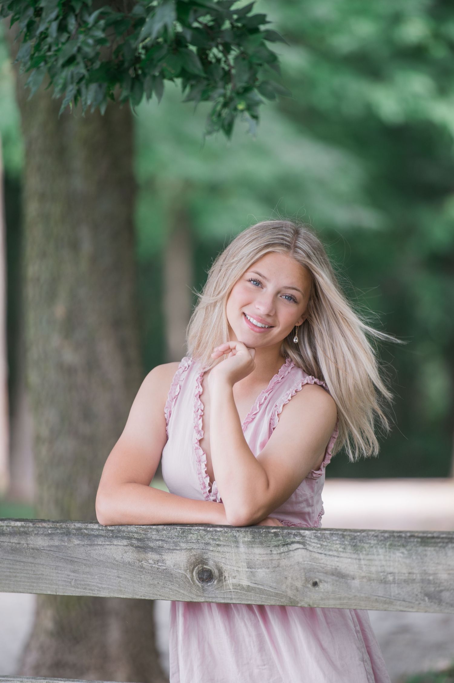 A senior portrait session Dublin, Ohio in a pink dress poses casually on a rustic wooden fence surrounded by green summer foliage.
