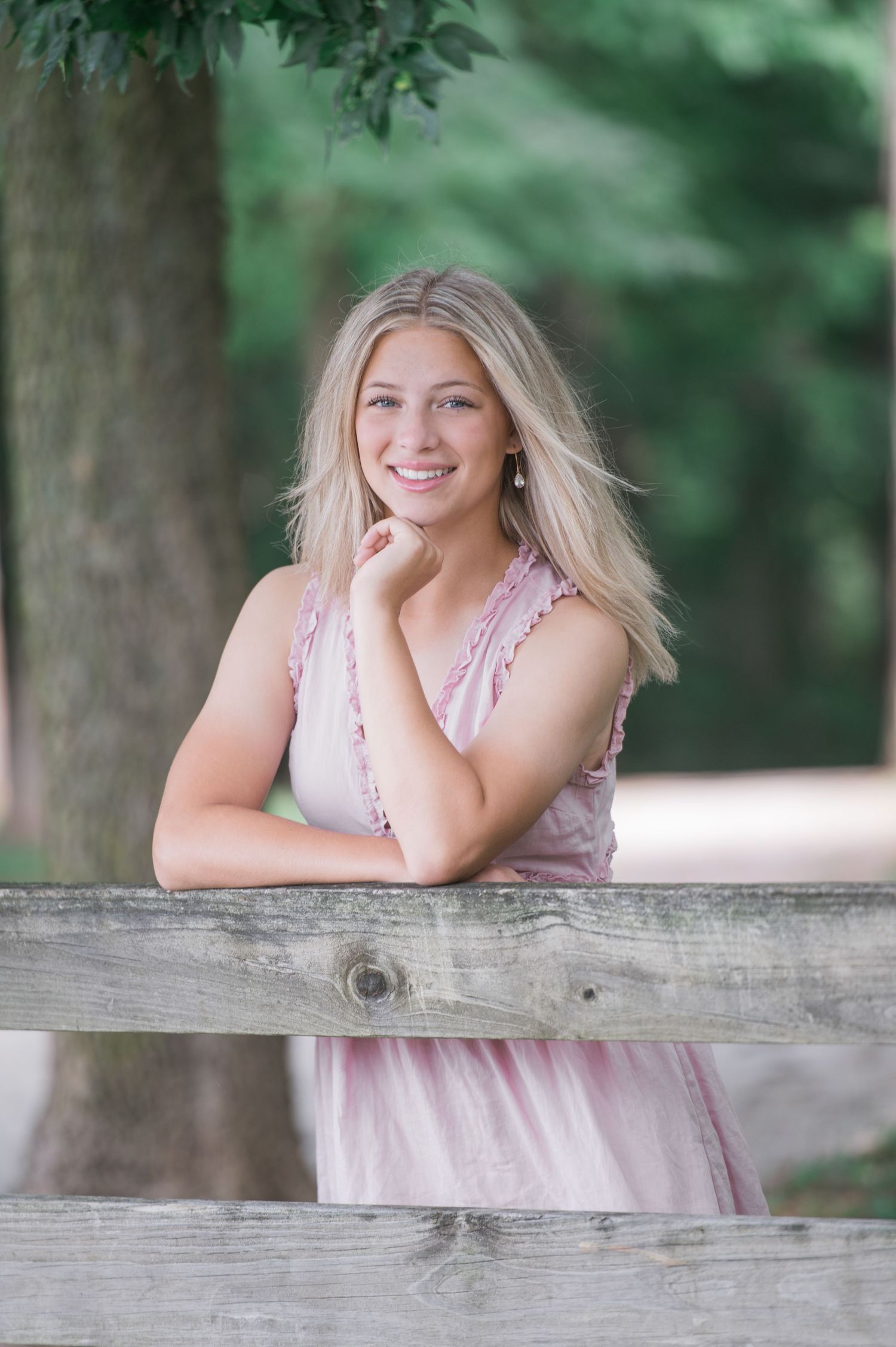 Outdoor senior portrait in Dublin, Ohio in a pink dress leaning on a rustic wooden fence in a park setting.