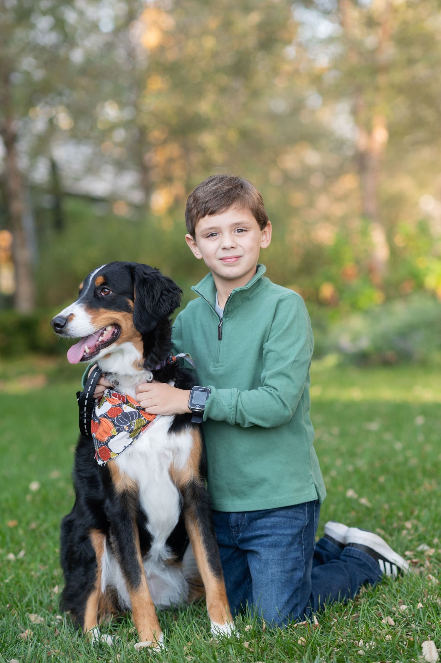 A young boy in a green pullover kneels on grass hugging a tri-color Bernese Mountain Dog wearing a bandana.
