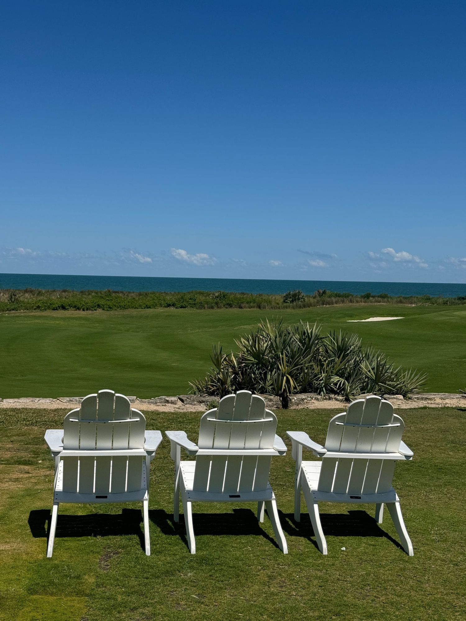 Three white Adirondack chairs overlook a lush golf course with ocean views under a clear blue sky.