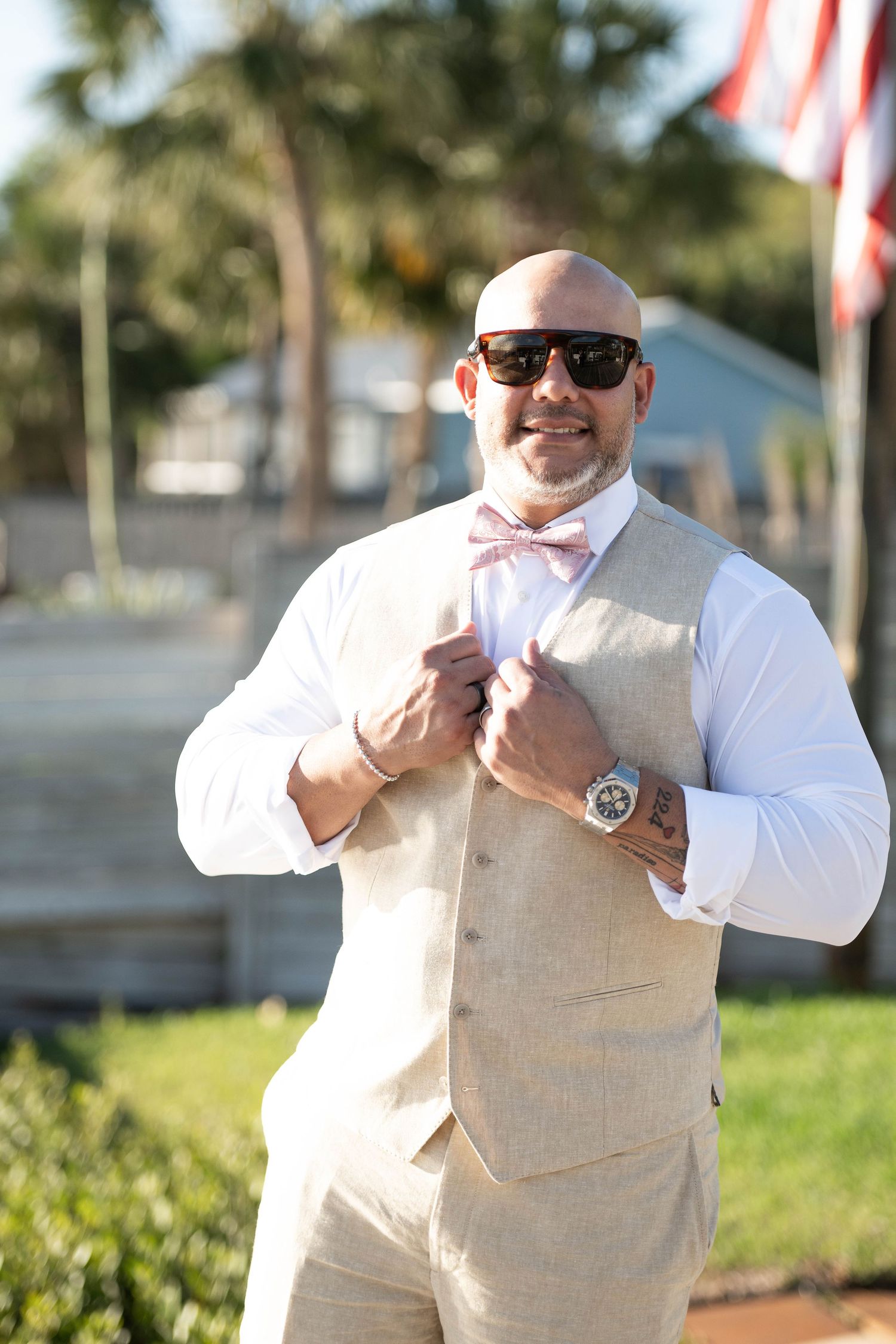 Bald groom in beige linen vest, pink bow tie, and sunglasses smiling outdoors at a sunny wedding venue.