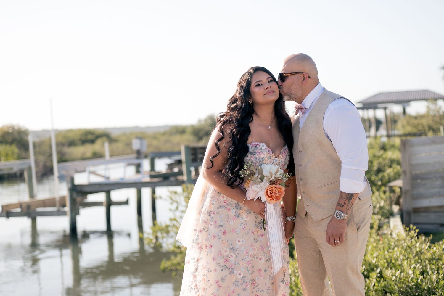 Bride in floral gown and groom in tan suit share a romantic moment by a waterfront dock on their wedding day.