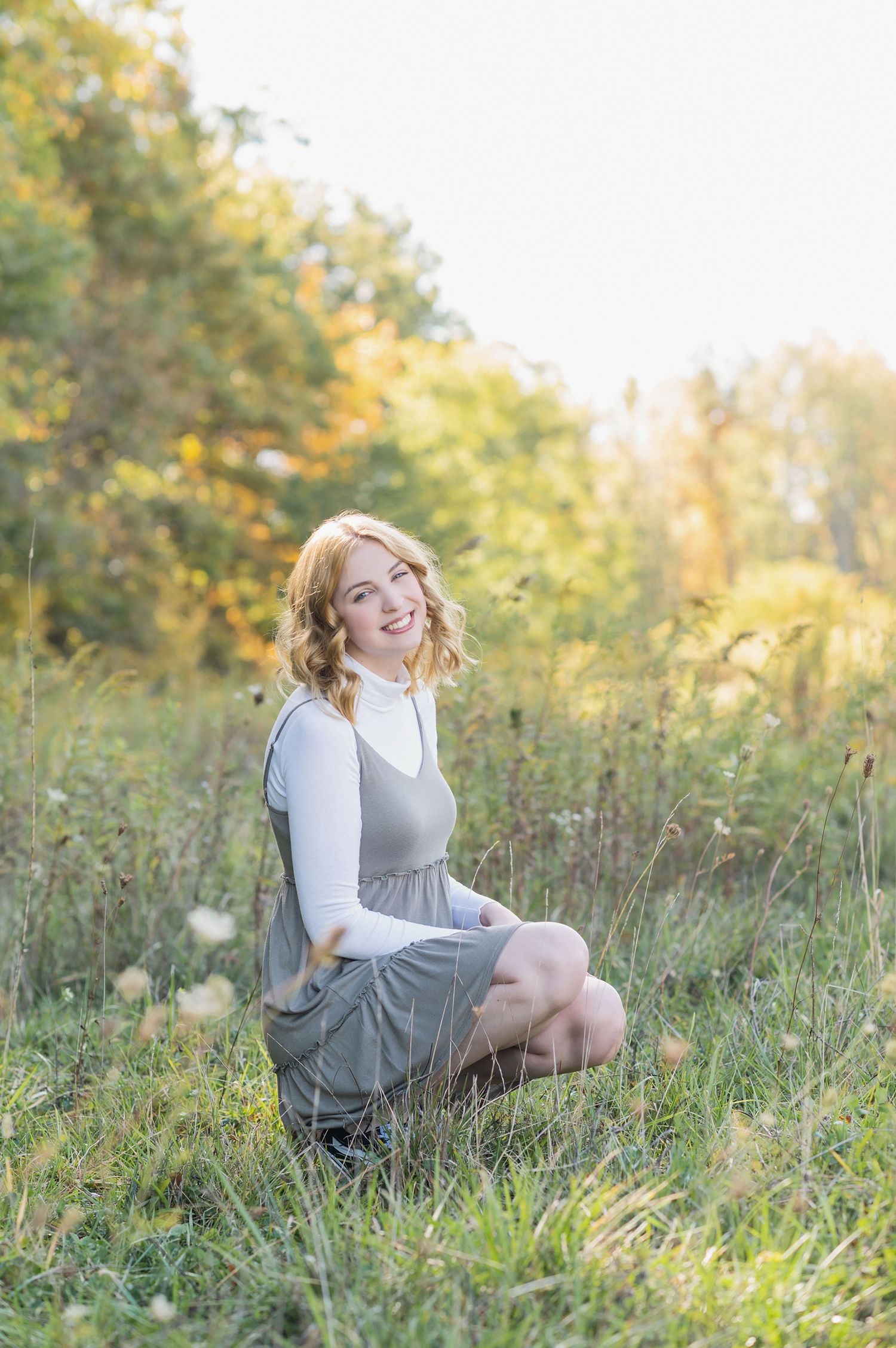A person in a gray dress and white shirt sits in a sunlit meadow with autumn foliage in the background.