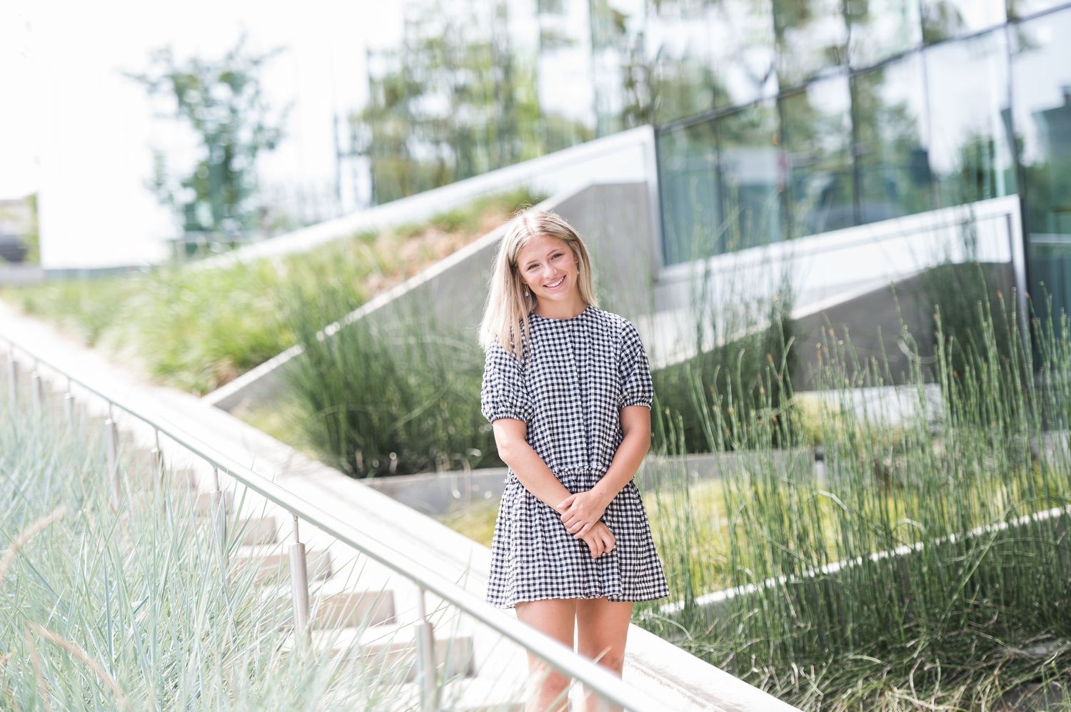 A Dublin Ohio Senior Portrait  in a checkered dress stands on outdoor steps surrounded by ornamental grasses on a sunny day.