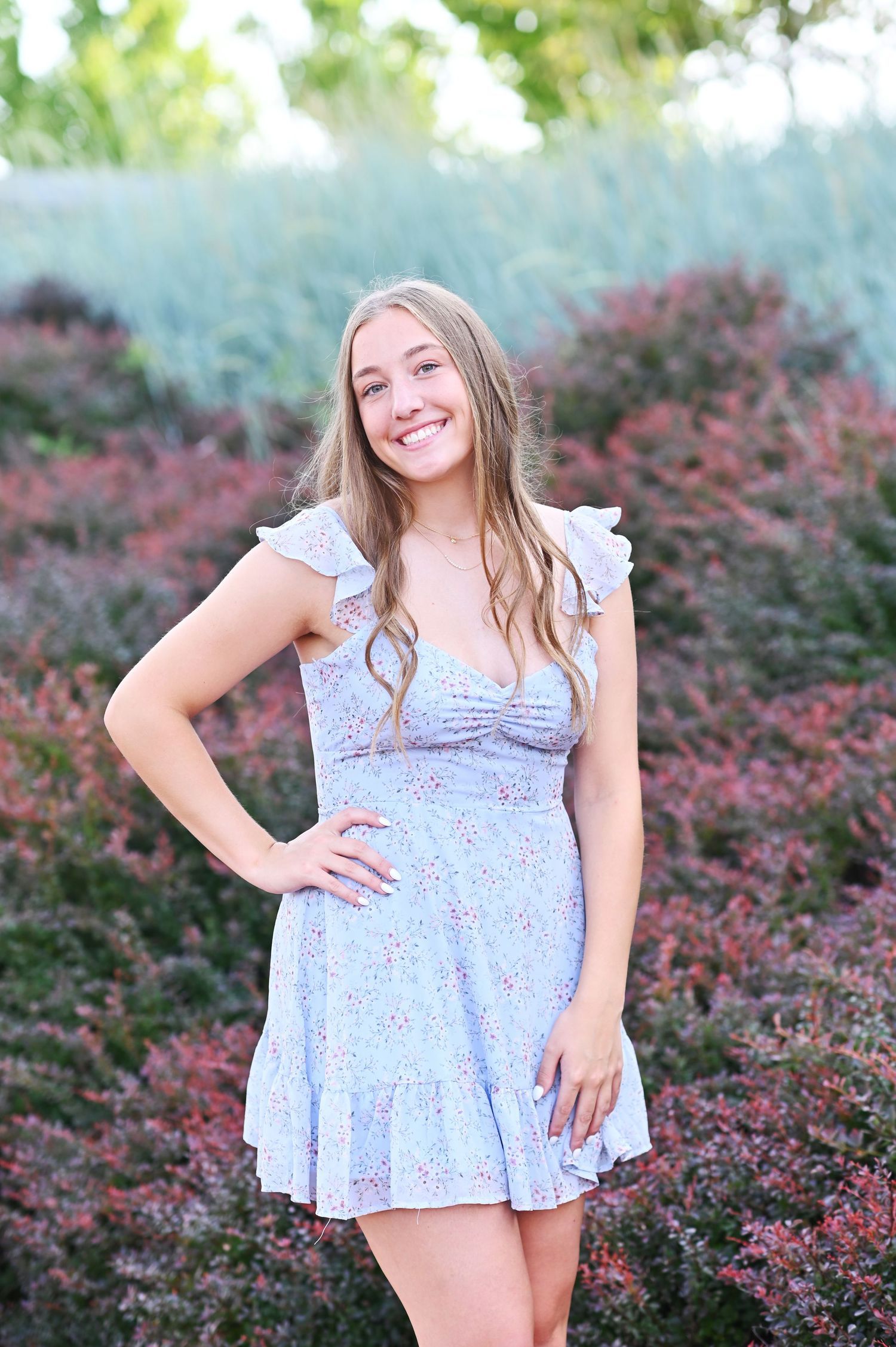 Person in light blue floral dress poses playfully among pink flowering bushes.
