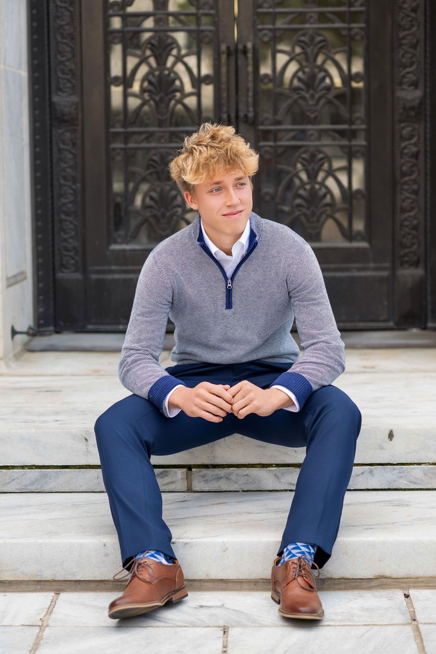 Young blond man in gray sweater and blue pants sitting on stone steps in front of ornate iron door.