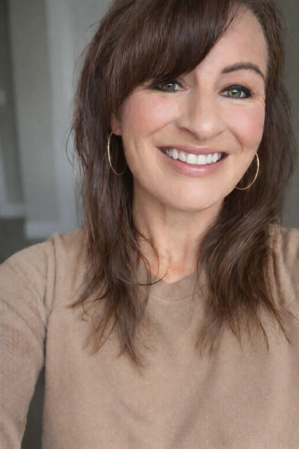 Smiling woman with brown hair, gold hoop earrings, and a beige sweater poses for a close-up portrait photo.