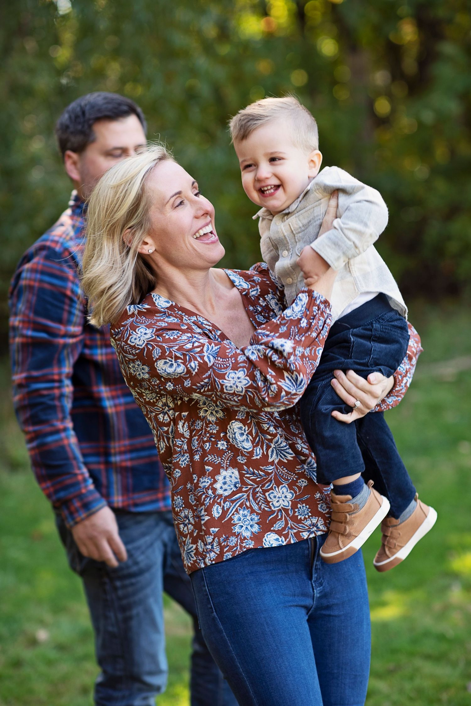 A joyful mother holds her laughing toddler outdoors while a man stands behind them in a lush green park setting.