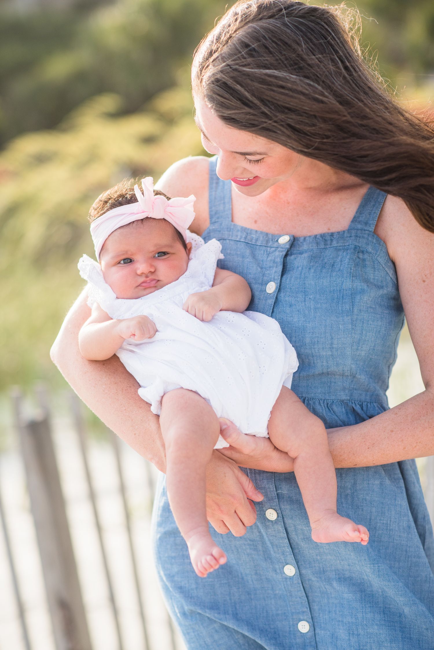 Young mother in blue dress lovingly holding her newborn baby with pink bow headband outdoors.