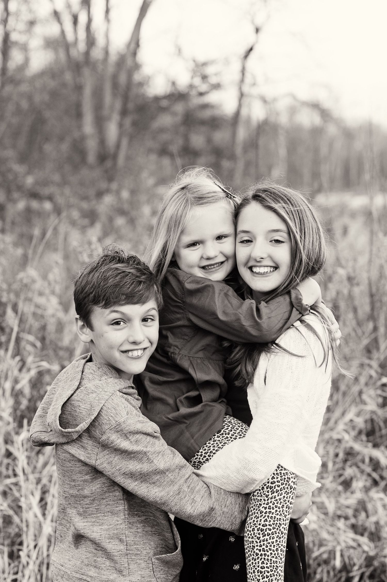 Black and white photo of three smiling children hugging outdoors in a field with trees in the background.