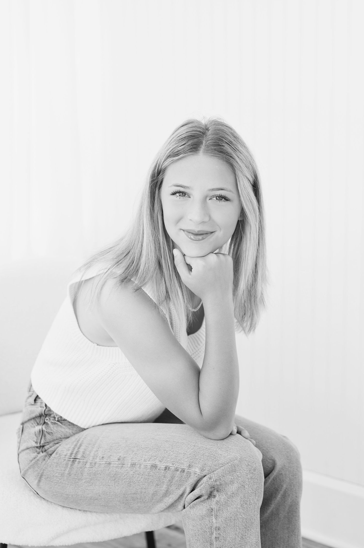 Black and white portrait of a smiling young blonde woman sitting relaxed with hand on chin, wearing jeans and a tank top.