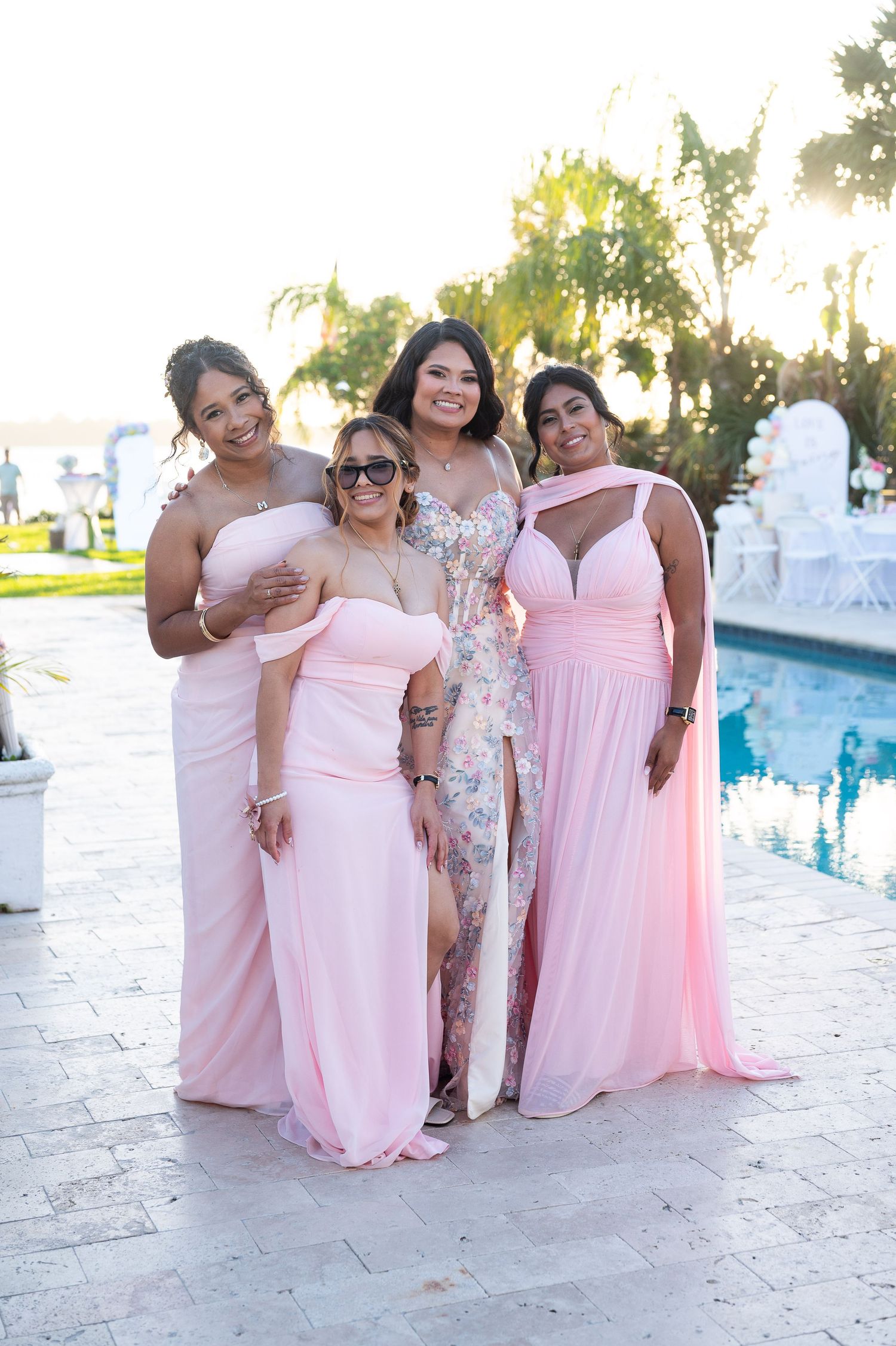 Four women in pink bridesmaid dresses pose together poolside at an outdoor wedding venue on a sunny day.