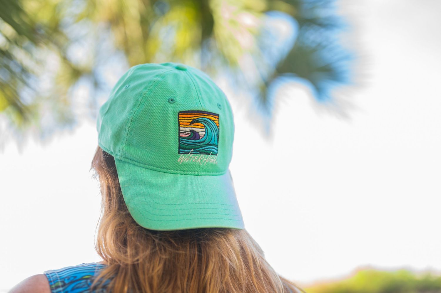 Person wearing a green baseball cap with a sunset logo stands in front of palm trees on a sunny day.