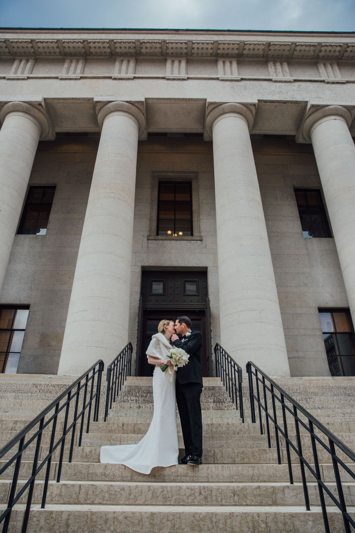 Wedding couple stands on steps of grand building with classical white columns.