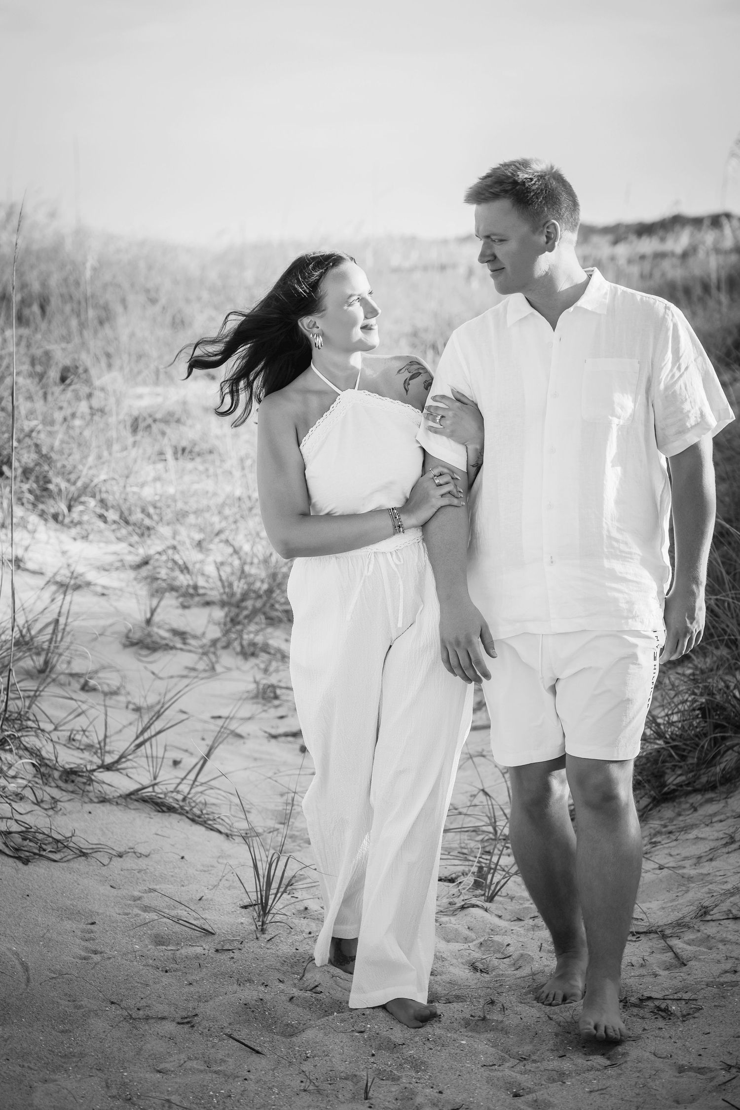 Black and white photo of a couple walking barefoot on sandy beach dunes, dressed in white outfits, smiling at each other.