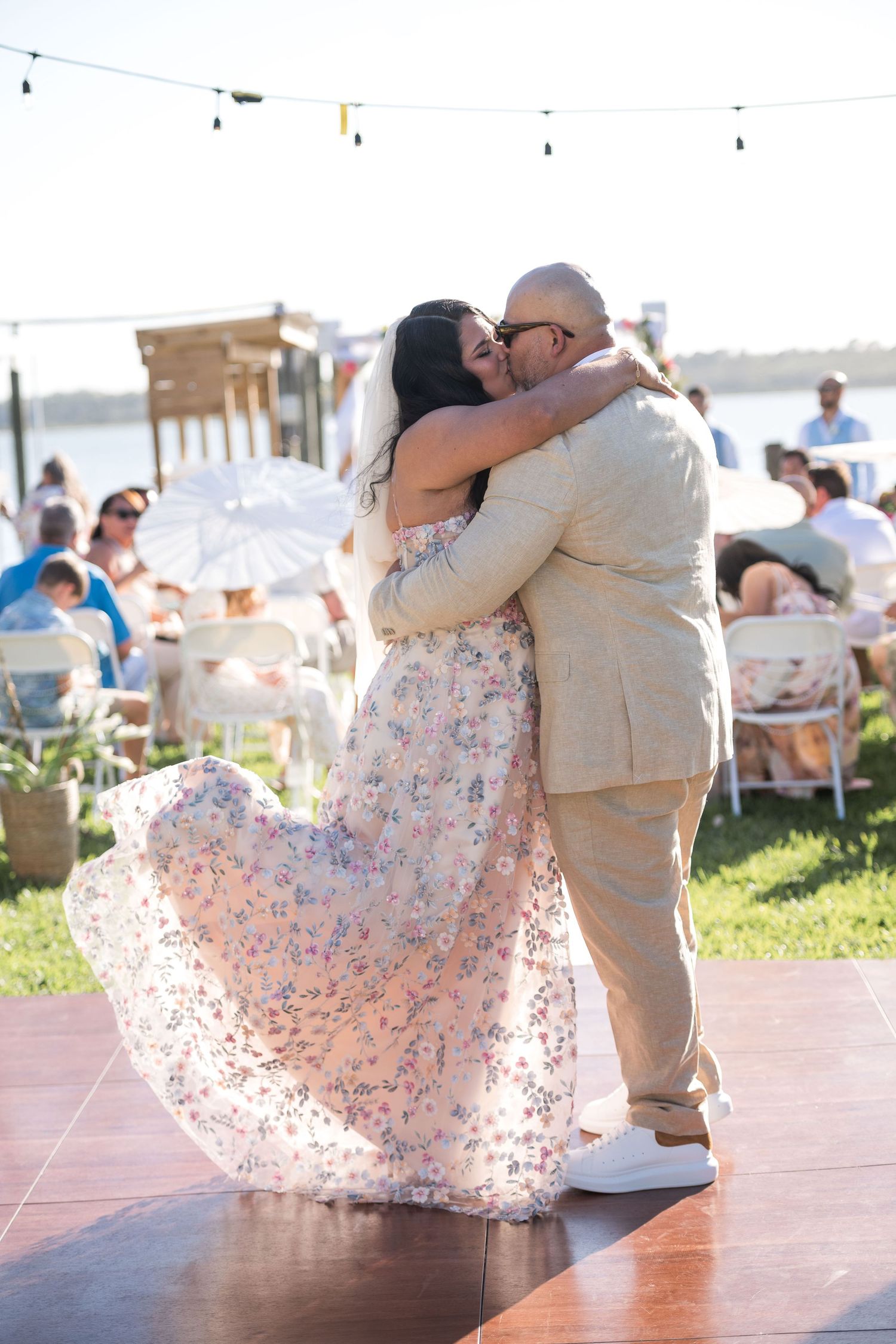 Couple sharing a first dance kiss in floral dress and tan suit at outdoor waterfront wedding reception.