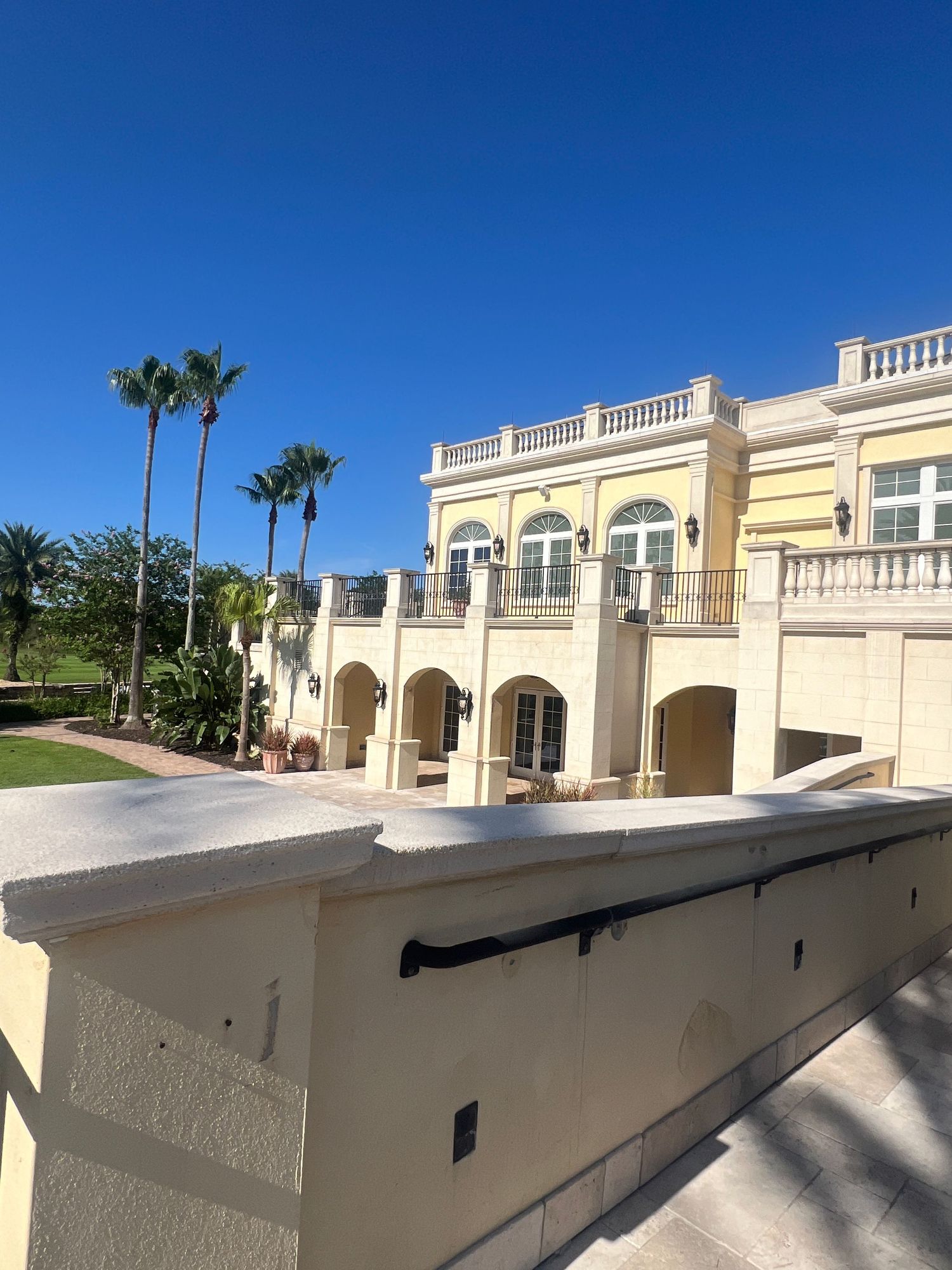 Elegant Mediterranean-style resort building with arched windows, white balustrades, and tall palm trees under a clear blue sky.