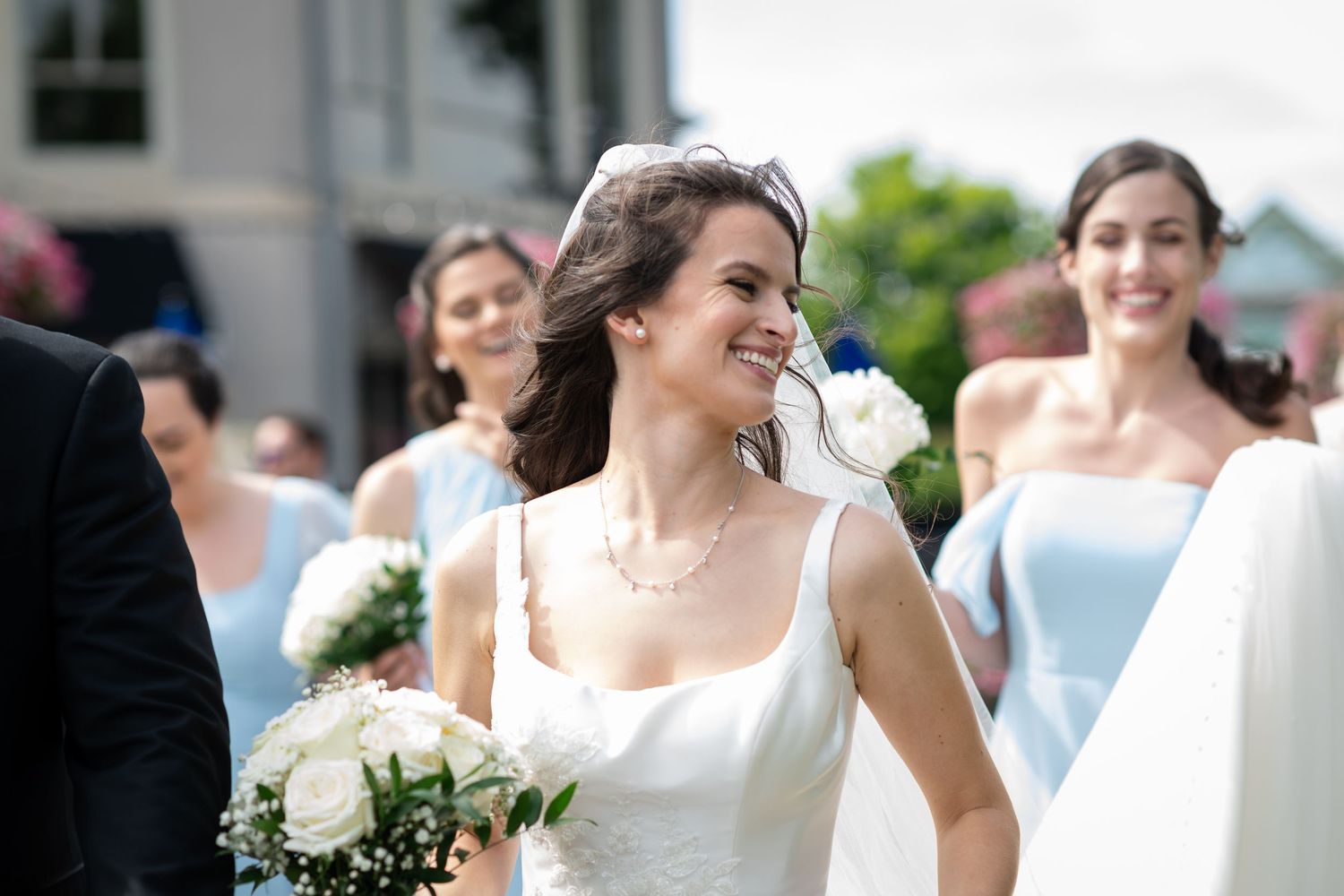 Smiling bride in white gown holding white bouquet walks with bridesmaids in light blue dresses at outdoor wedding.