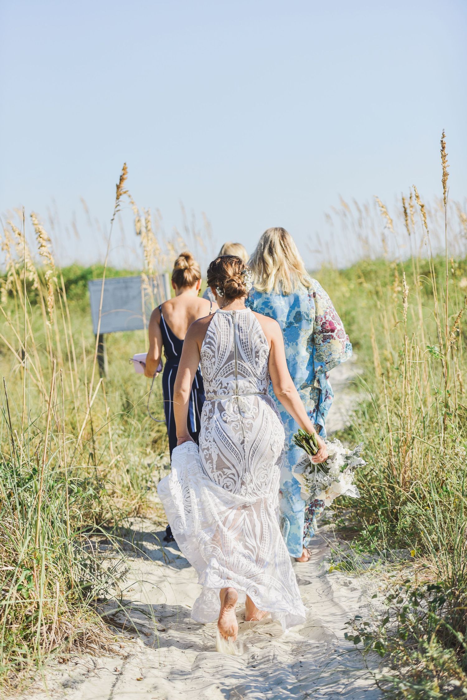 A group walks along a sandy beach path through sea oats and dune grass on a sunny summer day.