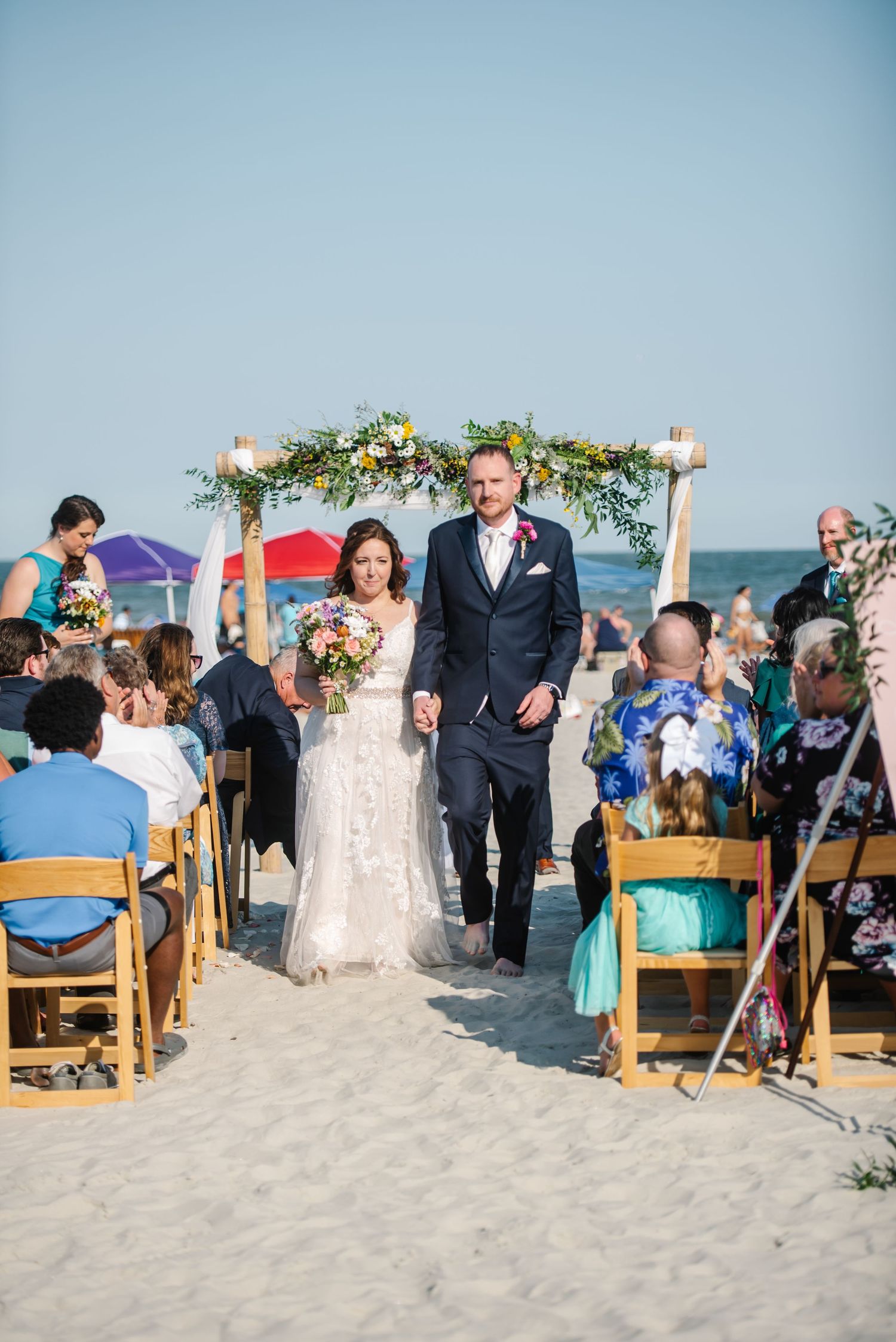 Beach wedding ceremony in progress with guests seated on wooden chairs facing decorated arch by the ocean. Beach Hilton Head