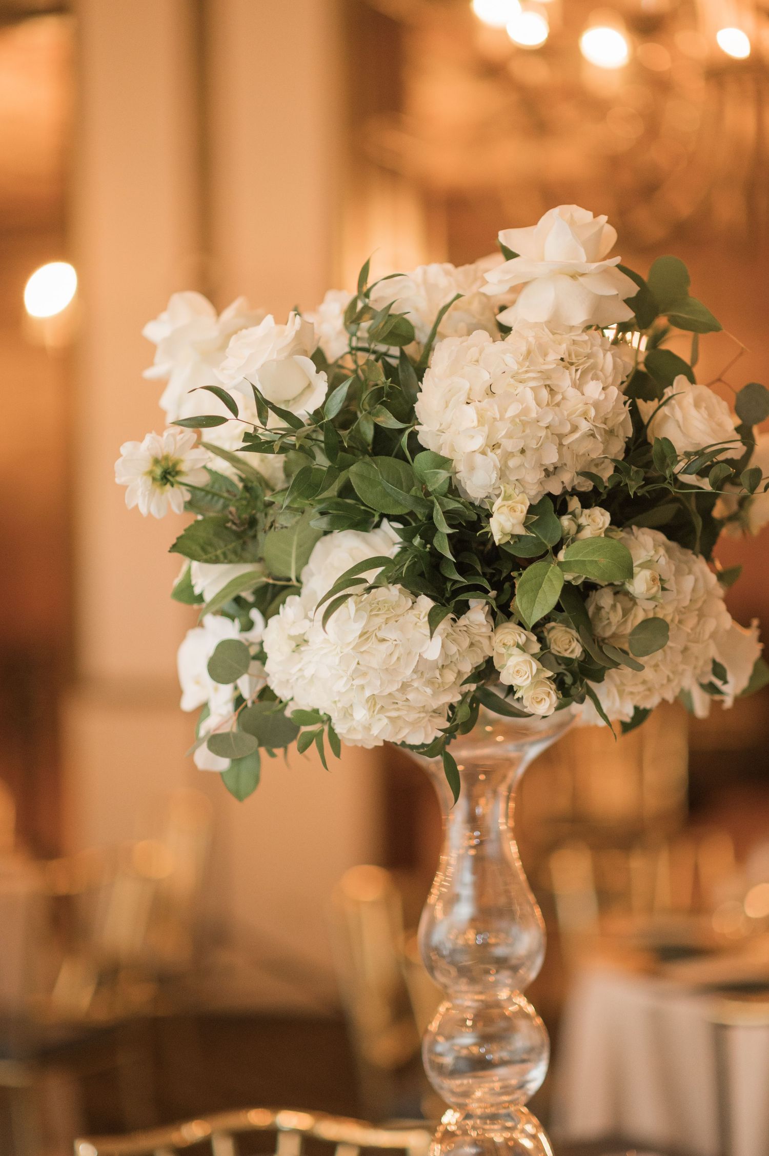 Wedding reception decor featuring white hydrangea and greenery centerpieces in glass vases with warm lighting.