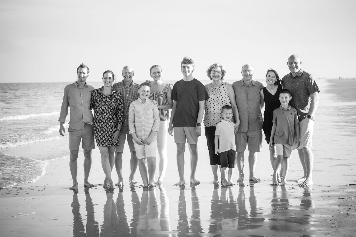 Extended family members pose together in black and white on a scenic beach shoreline.