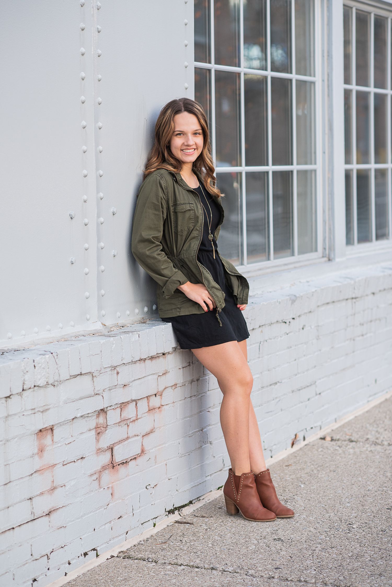 Someone poses by a white brick wall wearing boots and an olive green jacket.