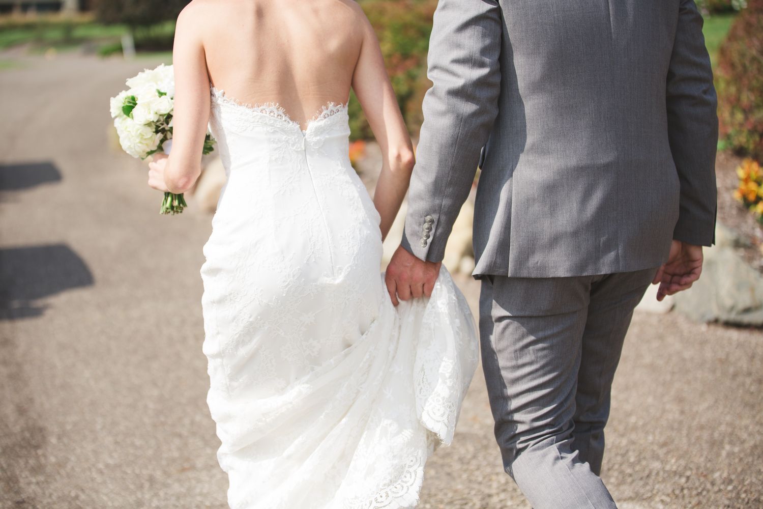 A bride in a white dress and groom in gray suit hold hands while walking down a garden path.