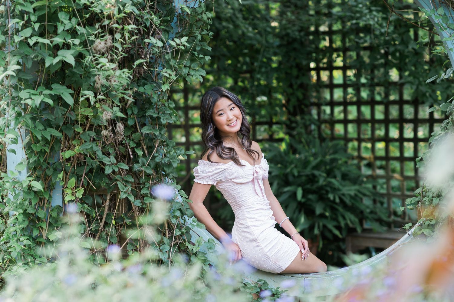 Young girl in a white dress poses playfully in a lush garden setting with green foliage and natural lighting.