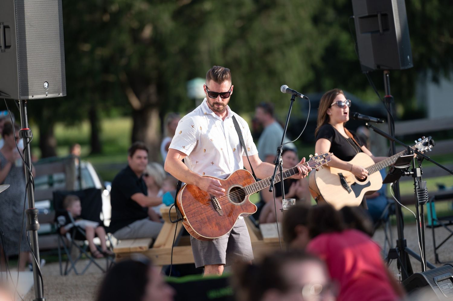 Musicians perform with acoustic guitars at an outdoor concert venue with audience members seated in the foreground.