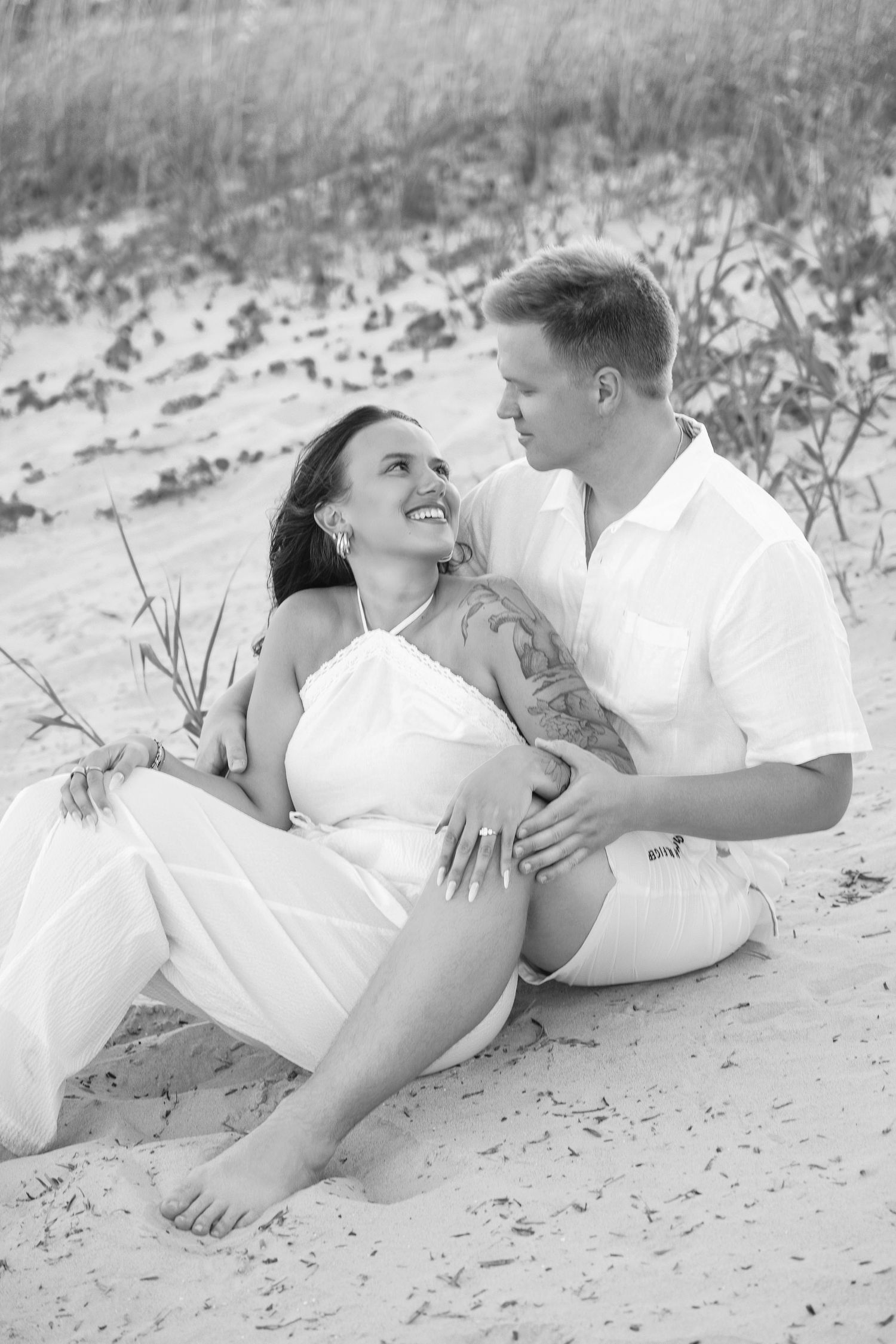 Black and white photo of a smiling couple sitting on beach sand, woman in white dress leaning against man in white shirt.