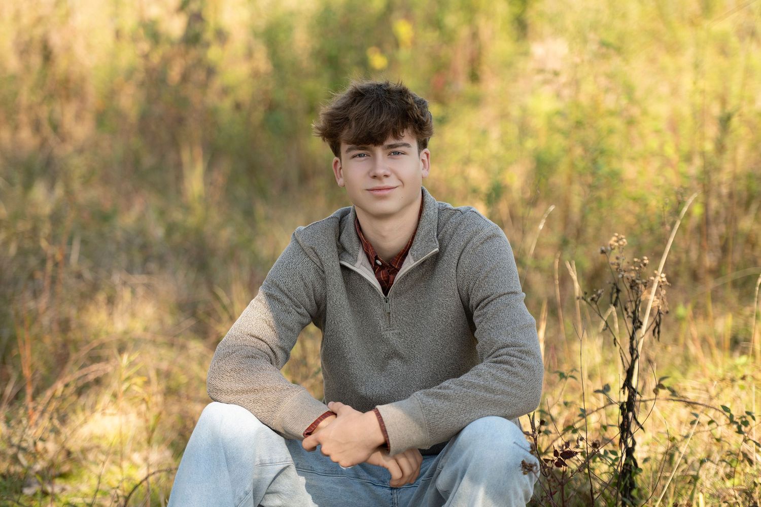 Young man in gray sweater sitting in a golden field, posing for a casual outdoor portrait photo.