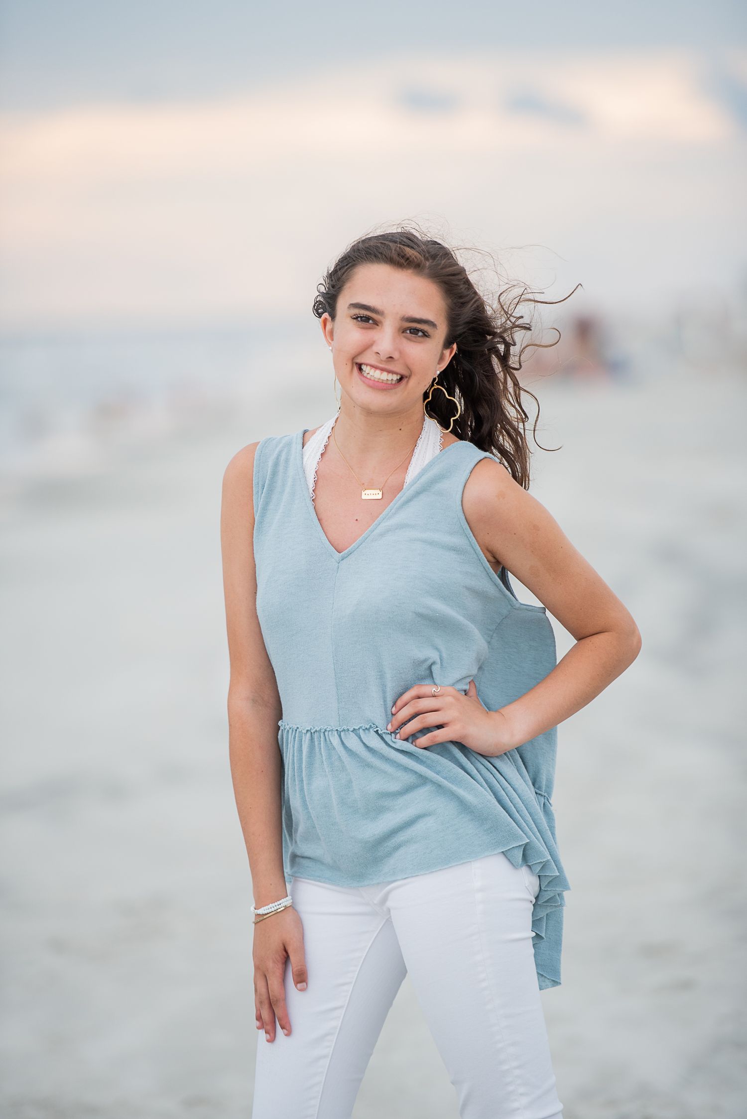 A person in a light blue sleeveless top and white pants poses on a beach at sunset with windswept hair.
