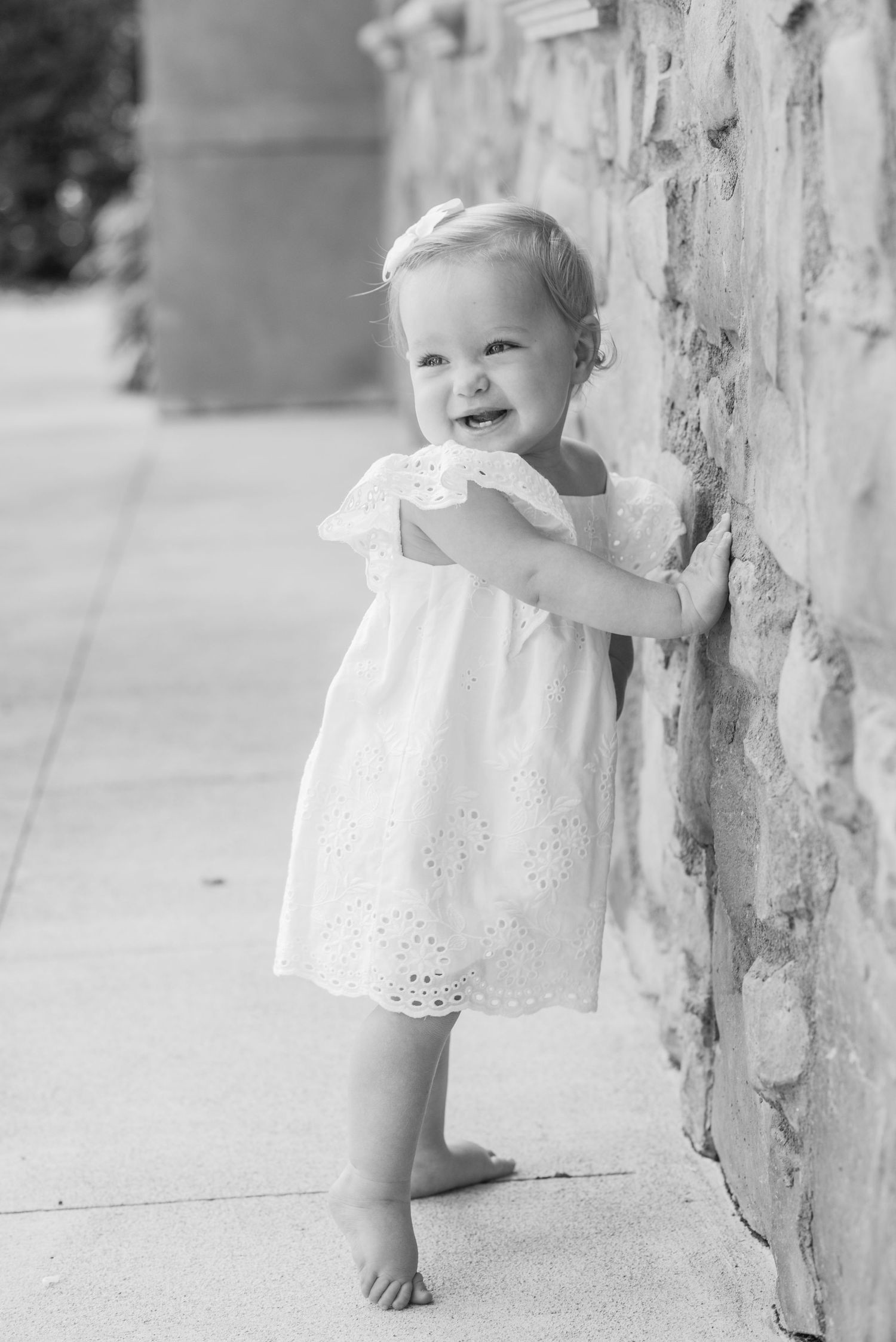 Black and white photo of a smiling toddler girl in a white eyelet dress standing barefoot against a stone wall.