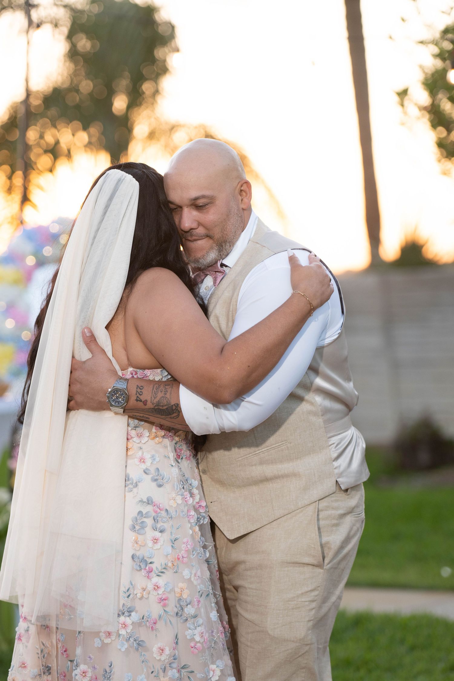 A bald groom in a tan vest embraces a bride in a floral dress and veil during an outdoor wedding ceremony.