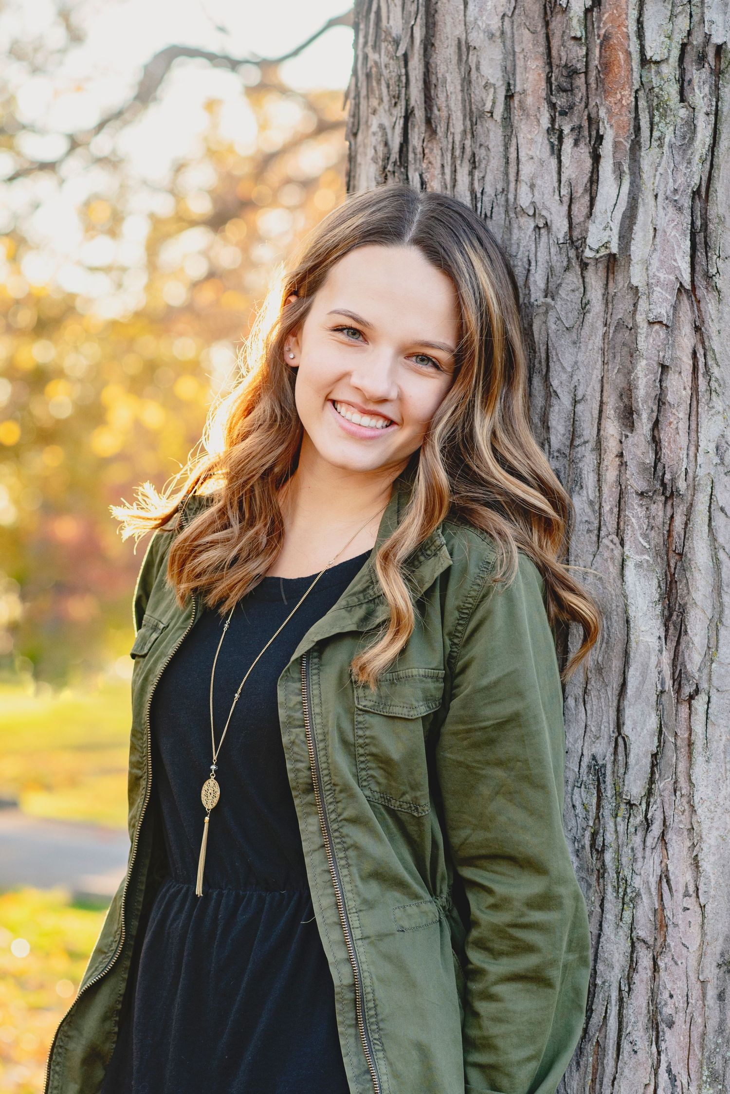 Autumn portrait against tree trunk wearing olive utility jacket and black dress with pendant necklace.