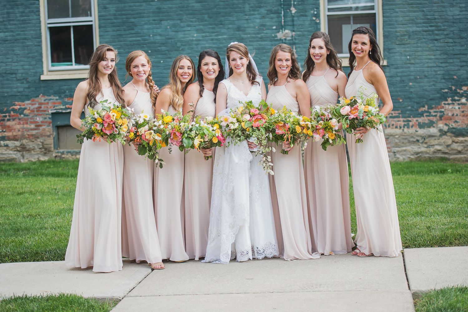 A row of bridesmaids in blush pink dresses stand together holding vibrant yellow sunflower bouquets.