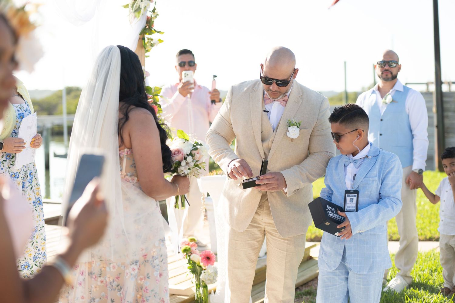 Groom in tan suit exchanges rings with bride in floral dress as young boy in blue suit watches at outdoor wedding ceremony.