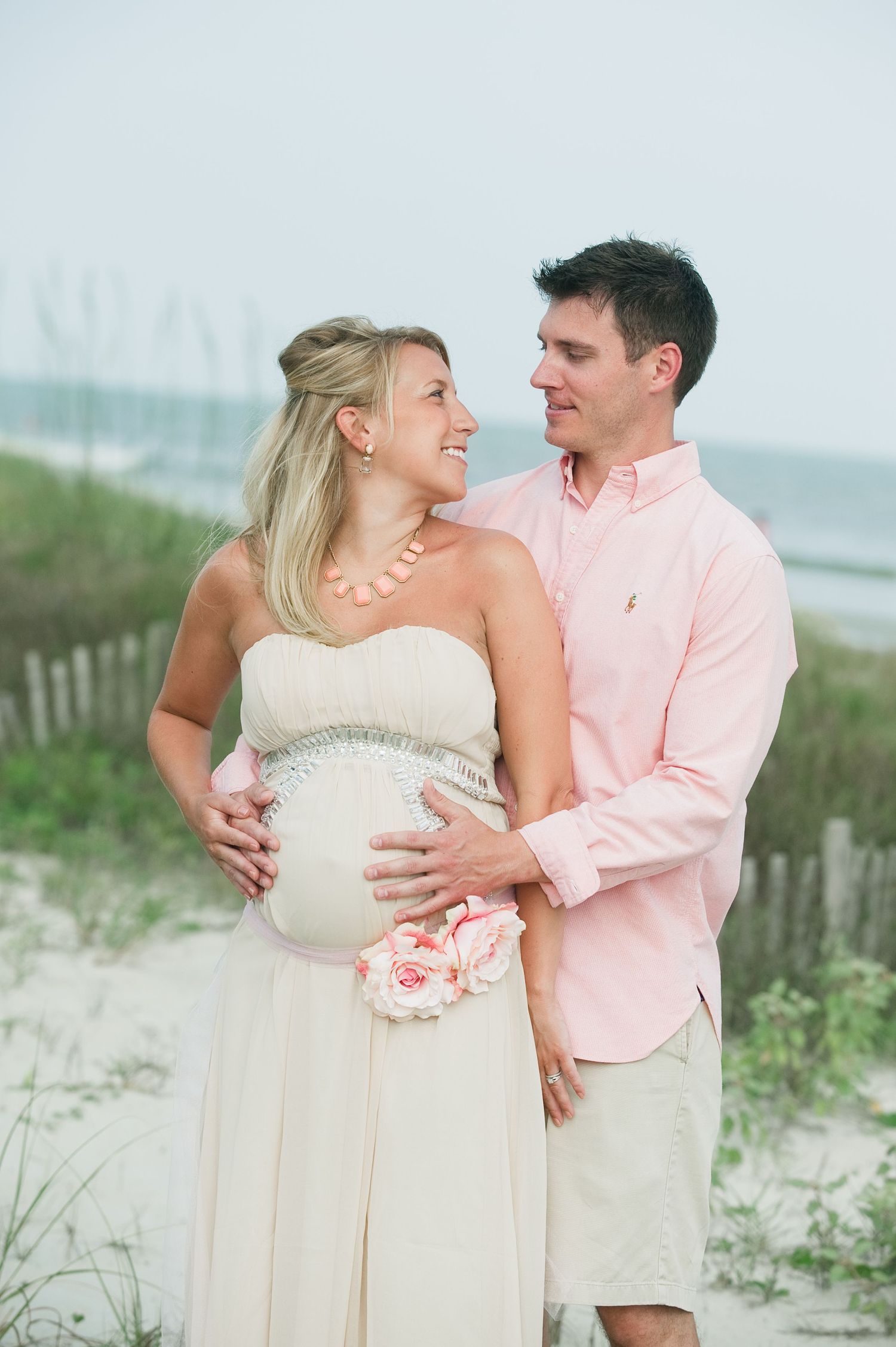 Pregnant woman in white dress with pink flowers and man in pink shirt embrace on a sandy beach.